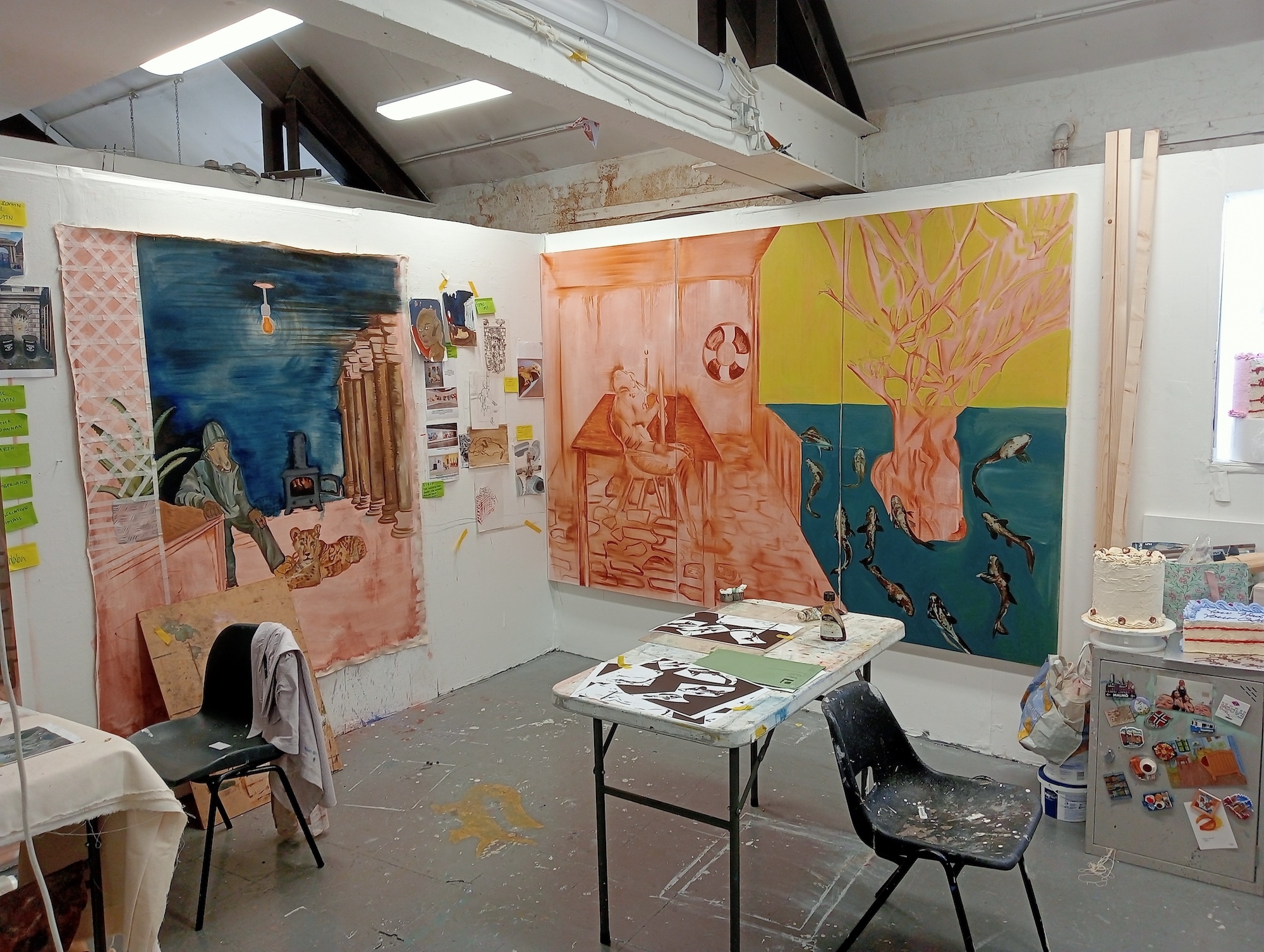 Art studio with large colourful paintings on walls featuring blue, orange, yellow and pink sections. White ceiling, grey floor, table with chairs and art supplies visible.