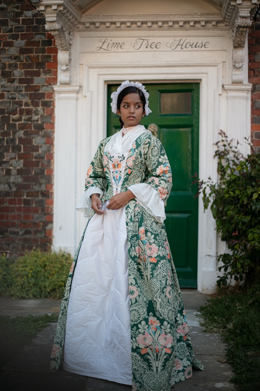 Woman in 18th-century dress with green floral pattern and white apron stands before Georgian brick house with green door and "Lime Tree House" sign.