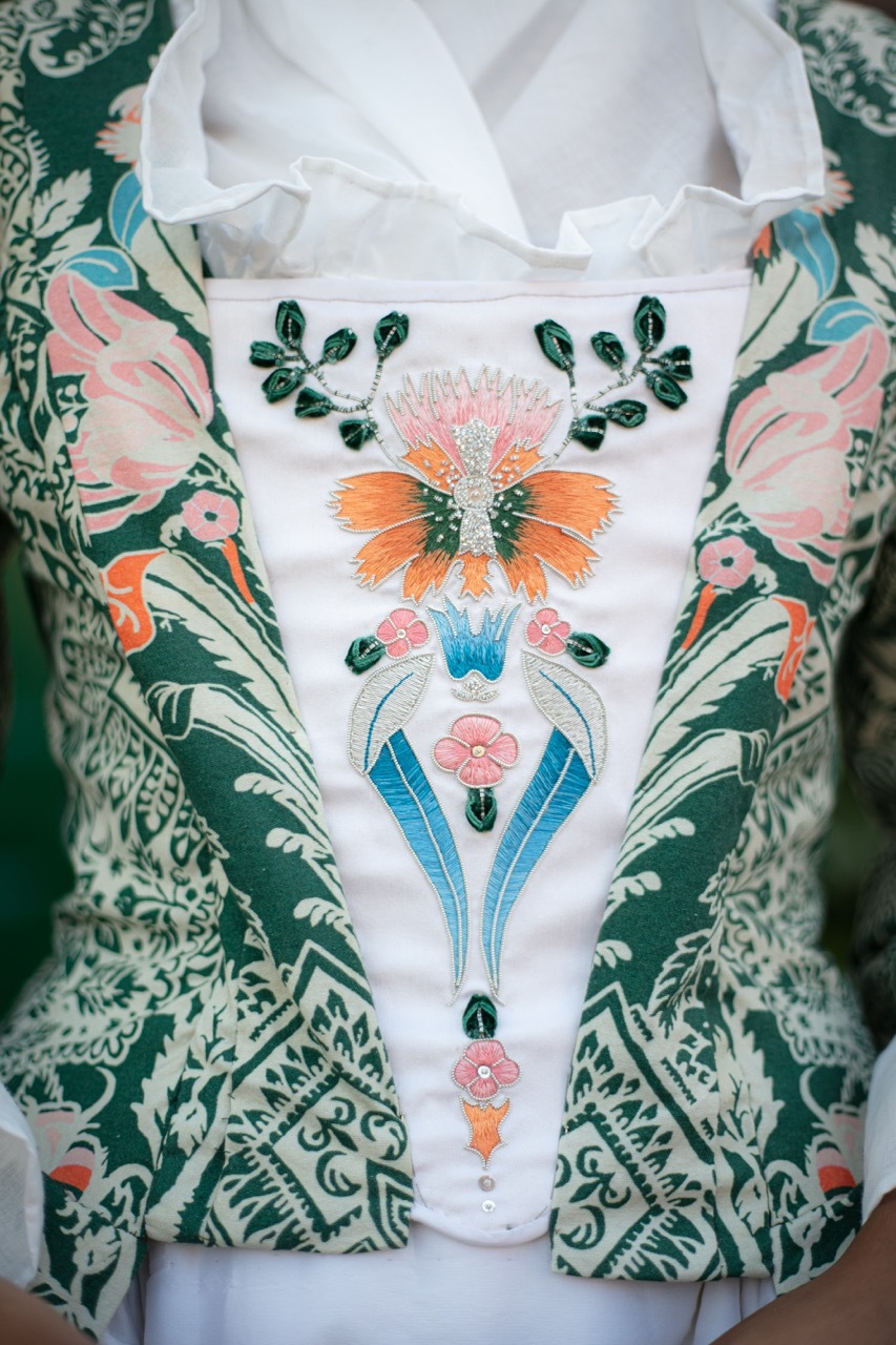 Traditional folk dress with white embroidered panel featuring orange flower, pink blossoms, green leaves, and blue stems on floral patterned fabric.