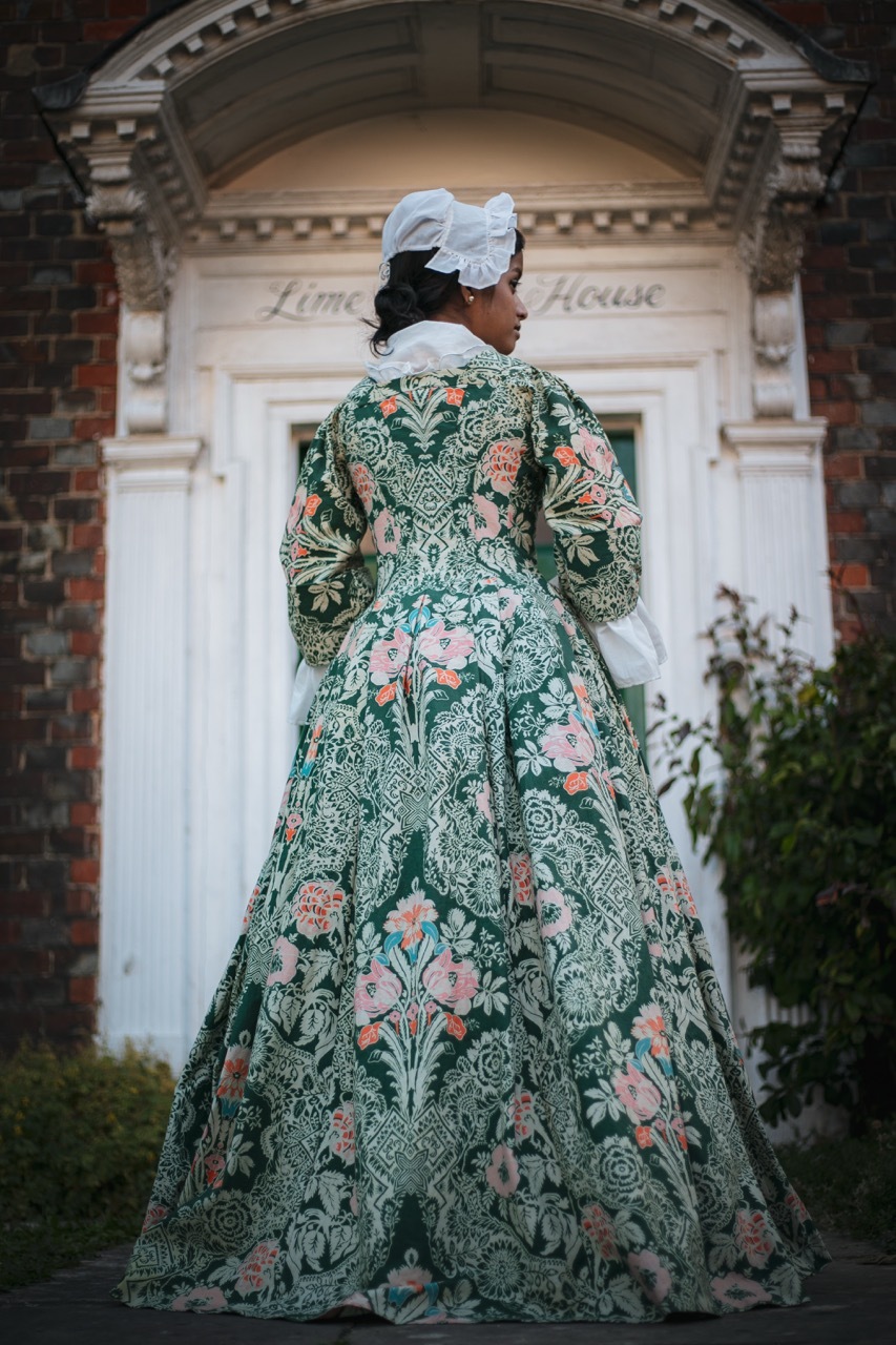 Woman in 18th-century green floral gown and white cap stands before Georgian doorway with classical pediment and brick facade.