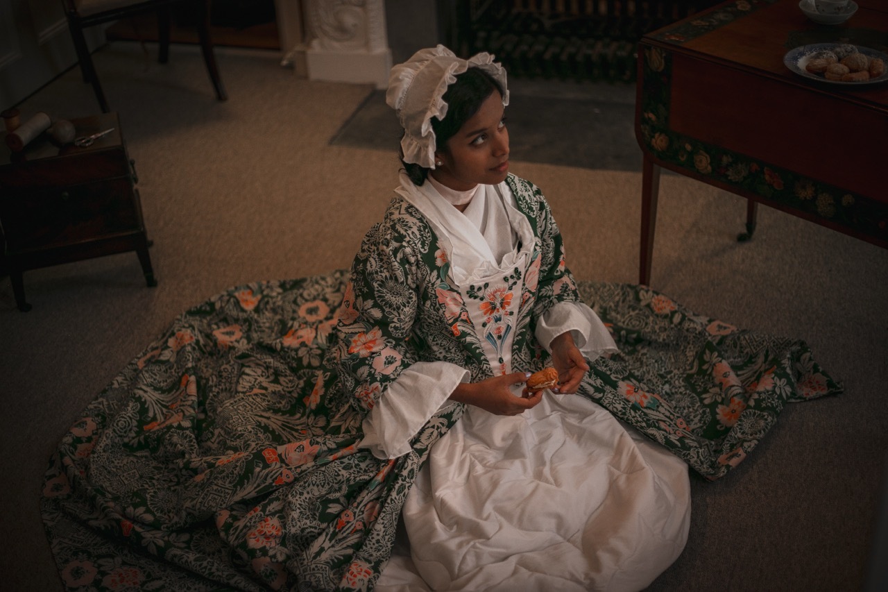 Woman in white dress with floral pattern sits on patterned fabric spread on ground, wearing white head covering with decorative elements.