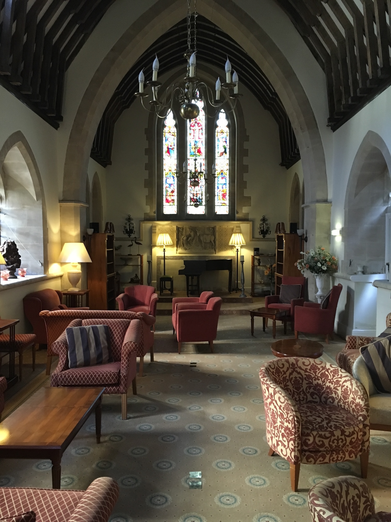 Gothic chapel interior with stone arches, exposed wooden beams, stained glass window, chandelier, red upholstered chairs and patterned floor