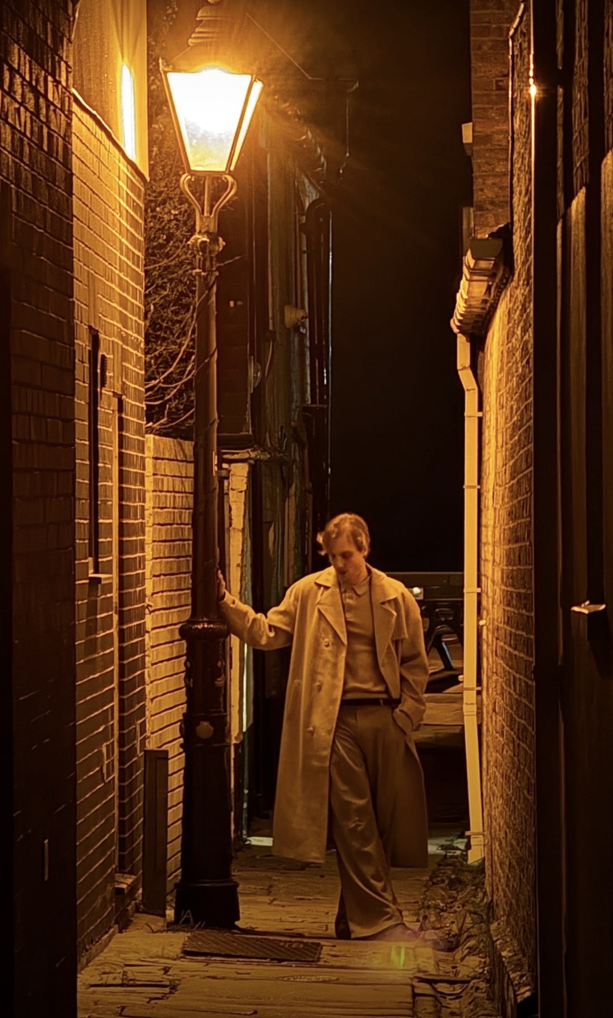A man in a beige trench coat stands in a narrow alley beside a lit streetlamp, with brick walls on both sides.