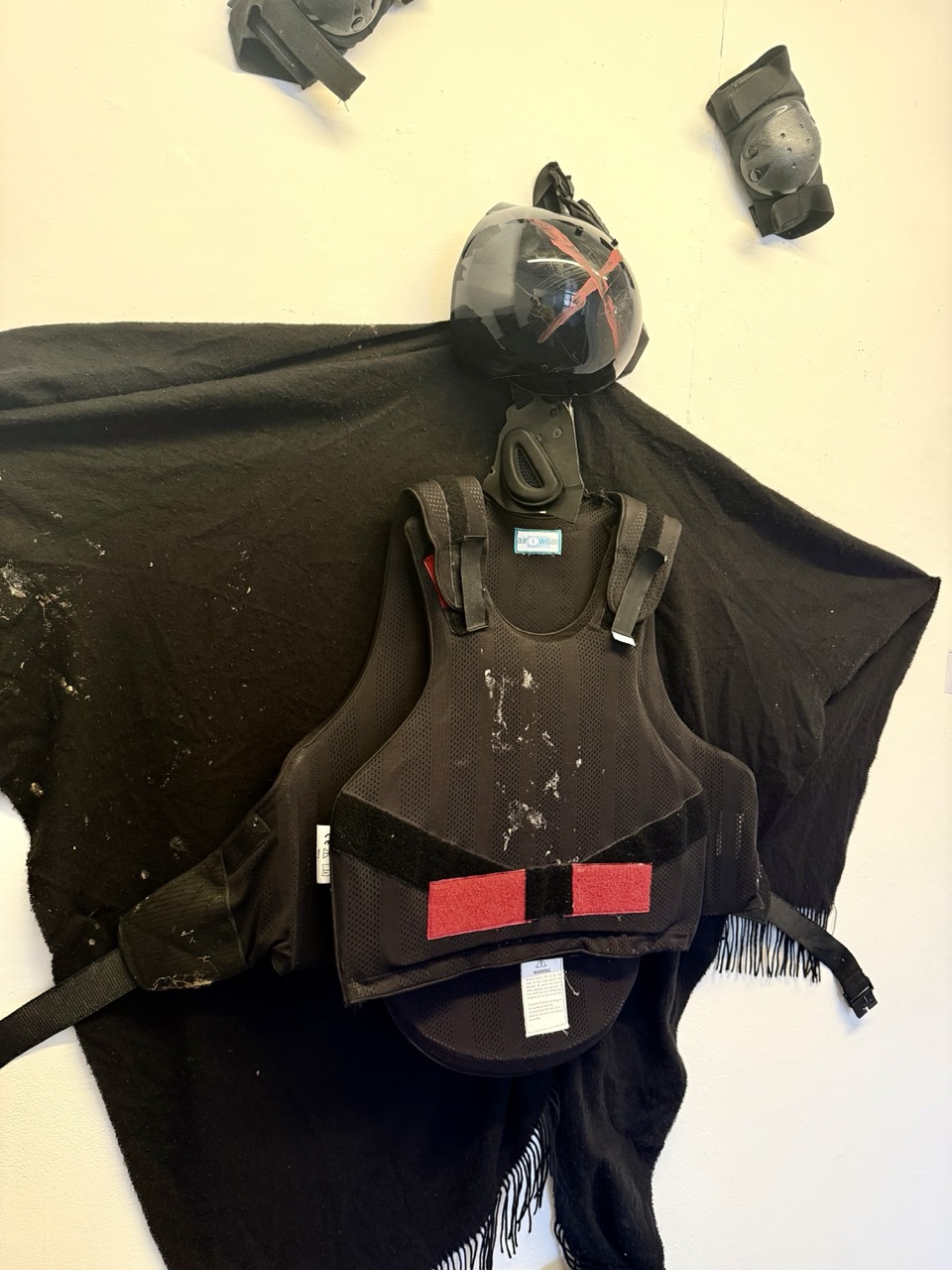 A black tactical vest with shoulder panels and a helmet mounted above it, displayed on a white wall with two protective items near the top corners.
