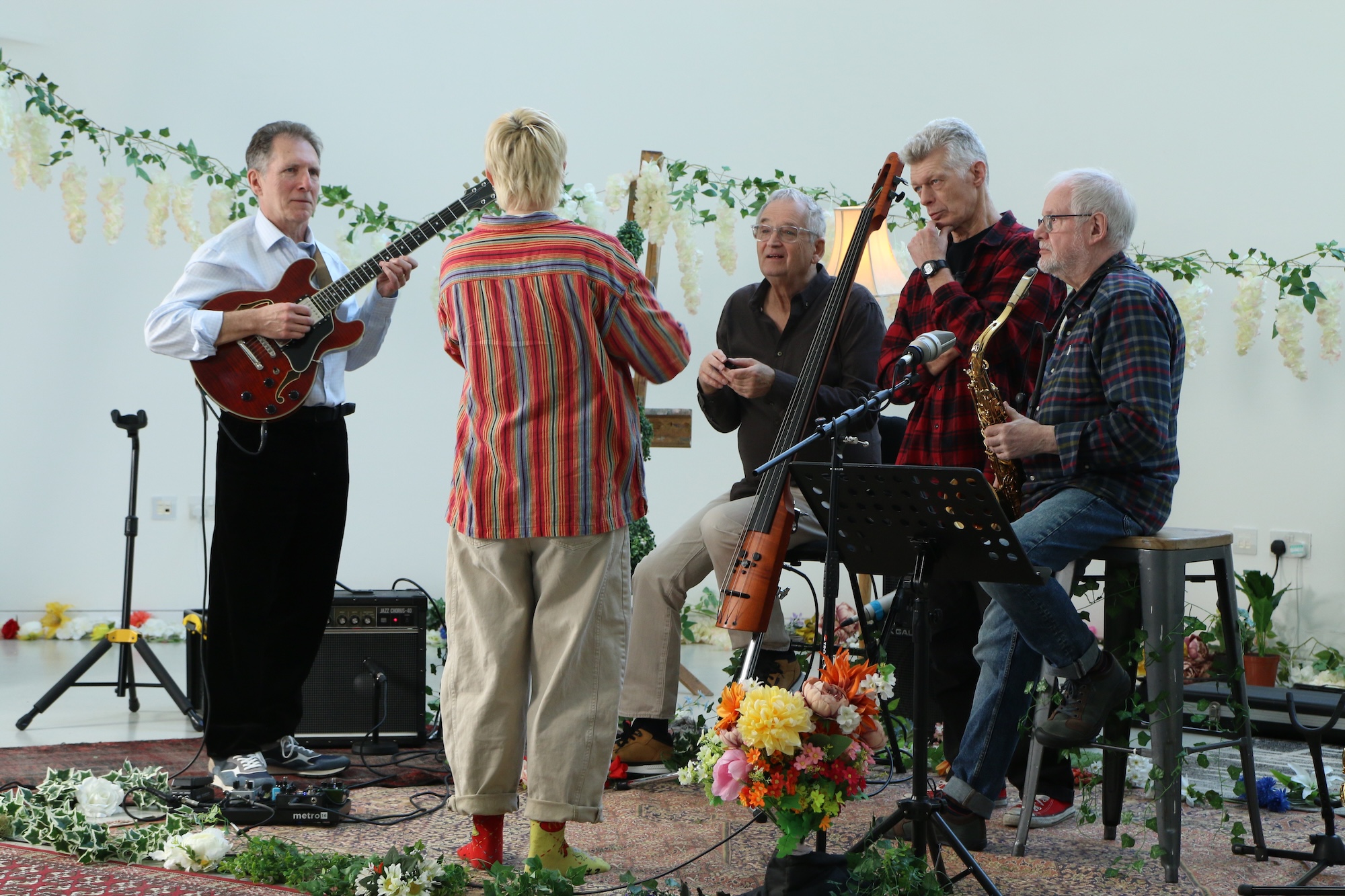A group of five older adults stands and sits with musical instruments in a bright room, with a guitar, bass and sheet music stand visible among floral decorations.