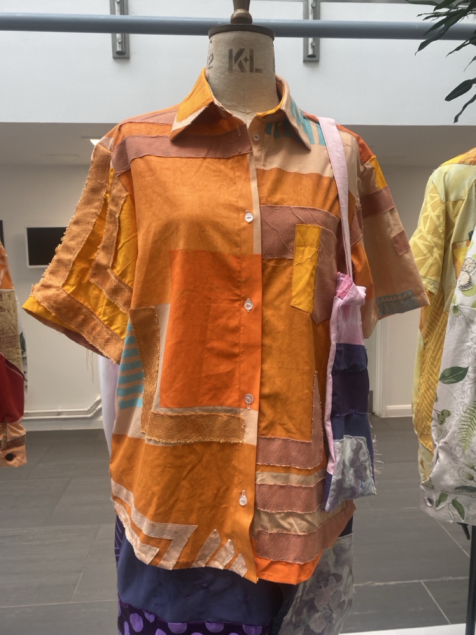 A brightly coloured short-sleeved shirt made of various patterned fabrics in shades of orange, peach, and tan, featuring a patch pocket and front buttons. The shirt is displayed on a mannequin in a well-lit space with other garments in the background.