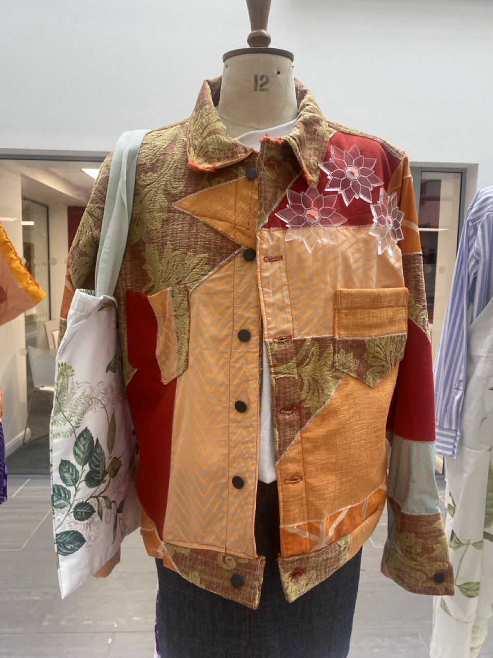 A multicoloured patchwork jacket displayed on a dress form. The jacket features various fabric textures and patterns including orange, red, and floral designs. A white tote bag with green leaf patterns hangs alongside.