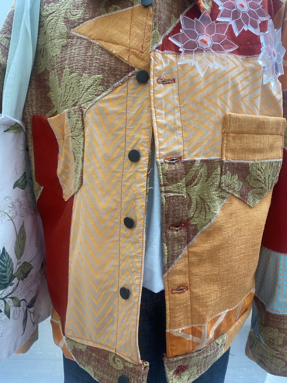 A close-up of a patchwork jacket featuring various fabrics in shades of orange, red, green, and white, with patterned sections including chevron and floral designs. The jacket has black buttons and a front pocket on the right side.