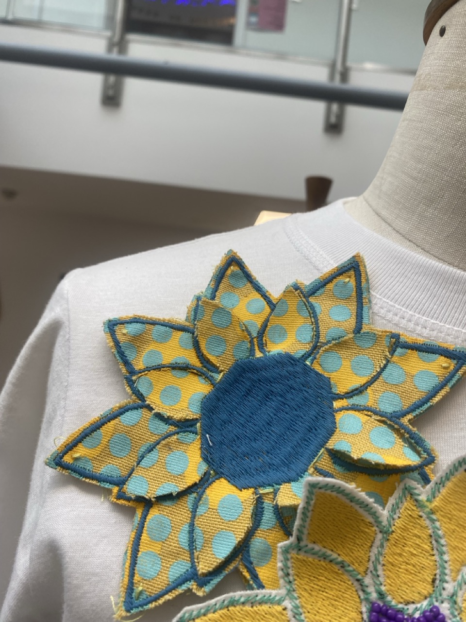 A close-up of a white garment featuring a large, colourful flower appliqué. The flower has turquoise petals with yellow polka dots, and a solid blue centre, displayed on a dressmaker's mannequin.