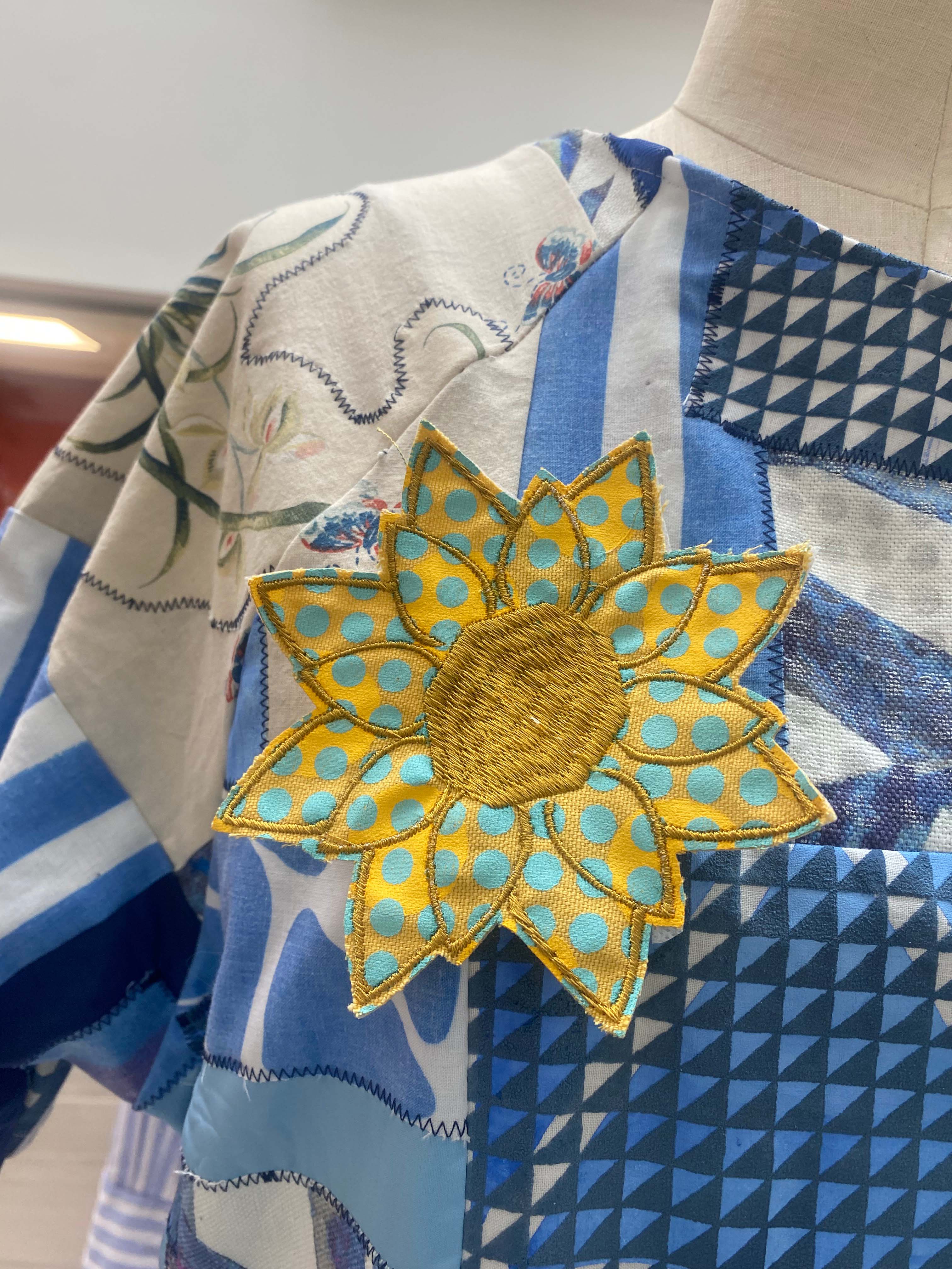A close-up of a mannequin wearing a multi-patterned shirt featuring geometric designs. Attached to the shirt is a large decorative flower brooch, with yellow petals dotted in blue and a brown centre.