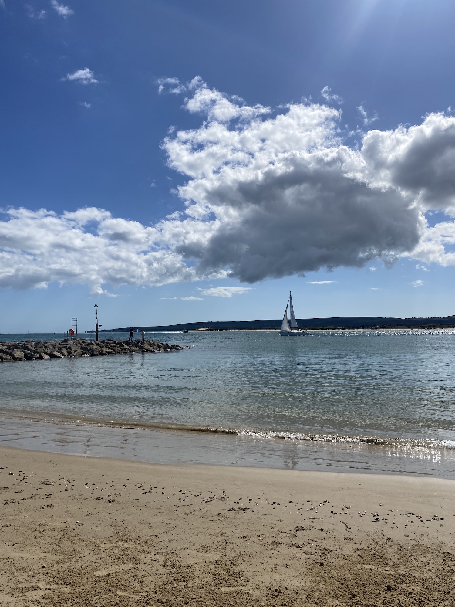 Beach and sea at Sandbanks under a cloudy sky with a small sailboat on the water in the distance.