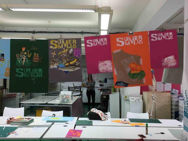A set of desks with drawing equipment in a drawing studio. Five pieces of coloured paper with text 'Summer Jam' along the top of each of them, hang above.