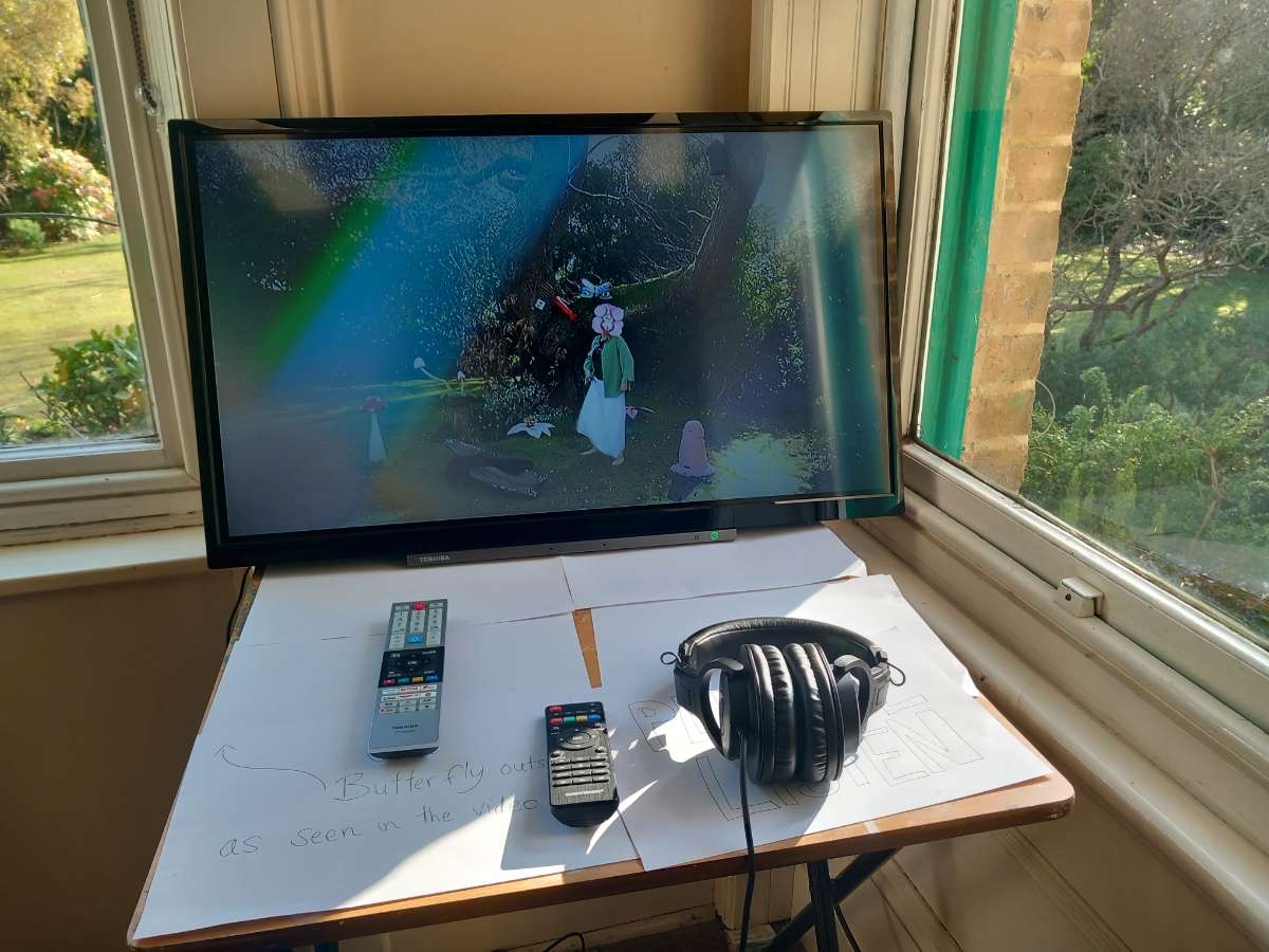 TV monitor screen showing abstract figure in garden, headphones and remote on table with paper in front of it, sunny garden windows to the side.