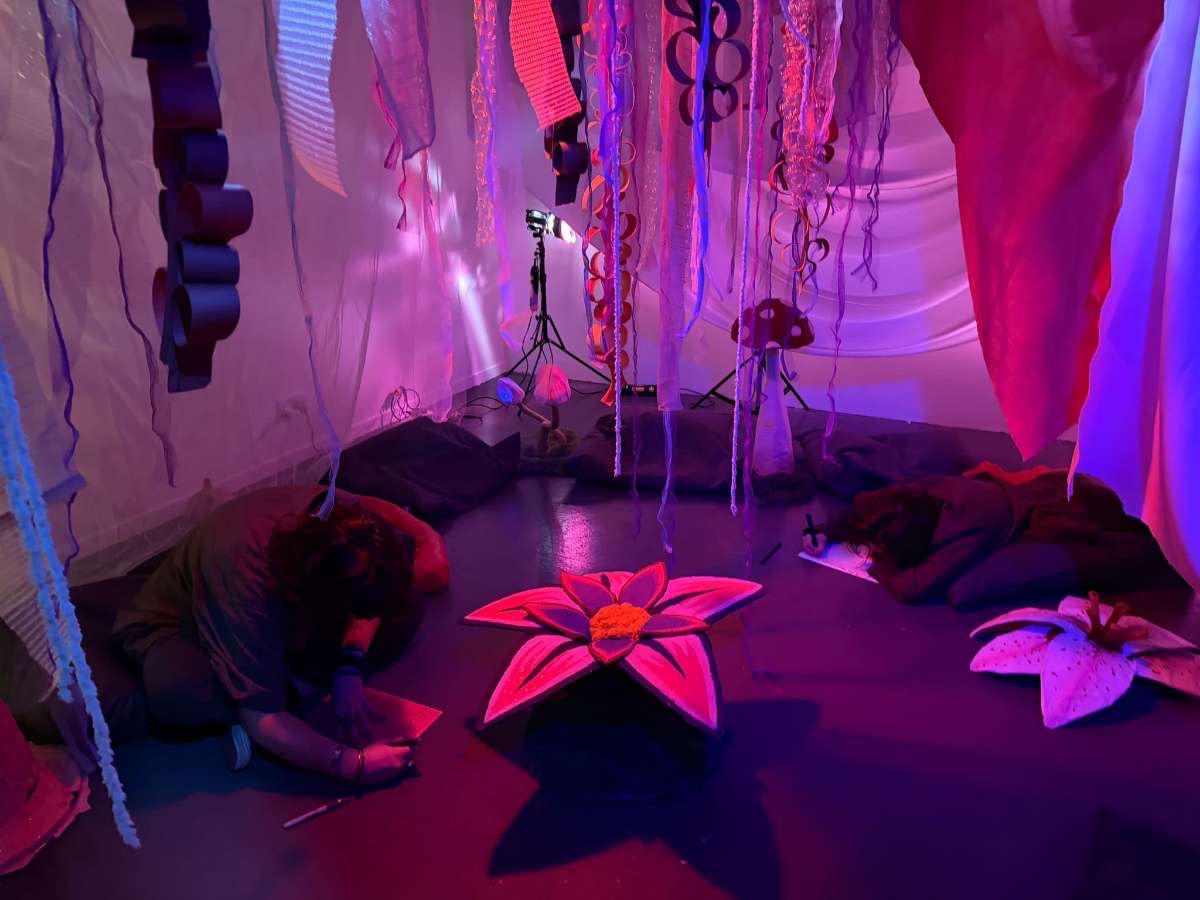 Abstract exhibition space with model flowers and mushrooms, purple, red and blue lighting, trailing fabrics from ceiling.