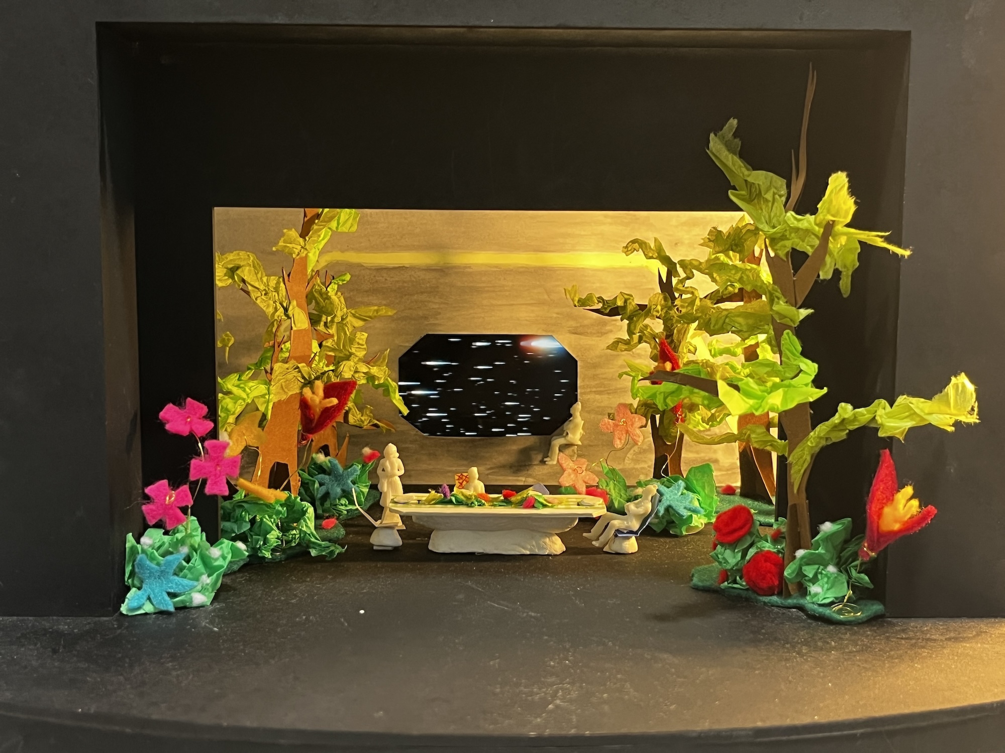 Model of a colourful theatre set with colourful plant life and a stone table in the centre. A wooden panel forms the background with an octagonal window and shooting stars visible outside.