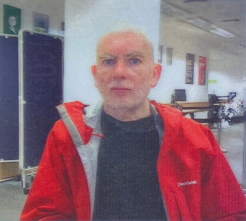 Bald man in red graduation gown with white hood and dark shirt, standing in office or classroom with desks and noticeboards.