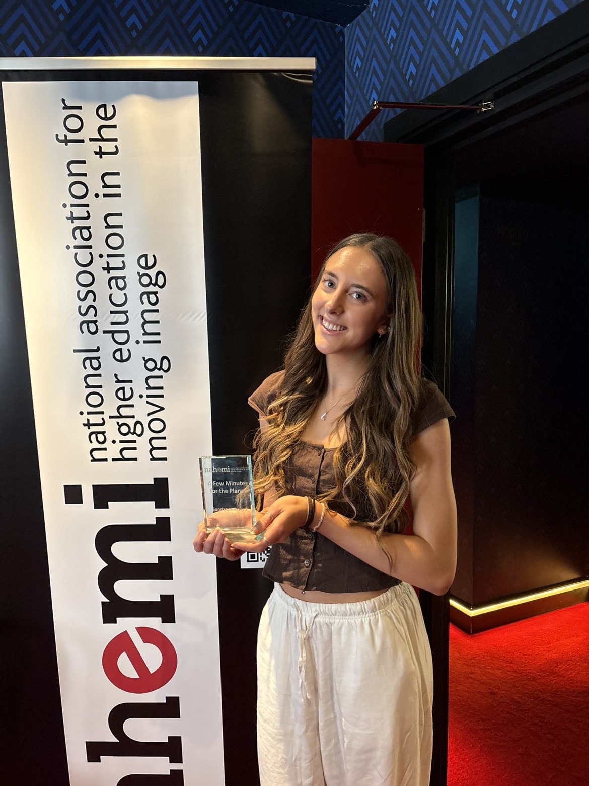 A young woman holds a clear award plaque beside a vertical banner for the national association for higher education moving image, in a dark exhibition space with red lighting.