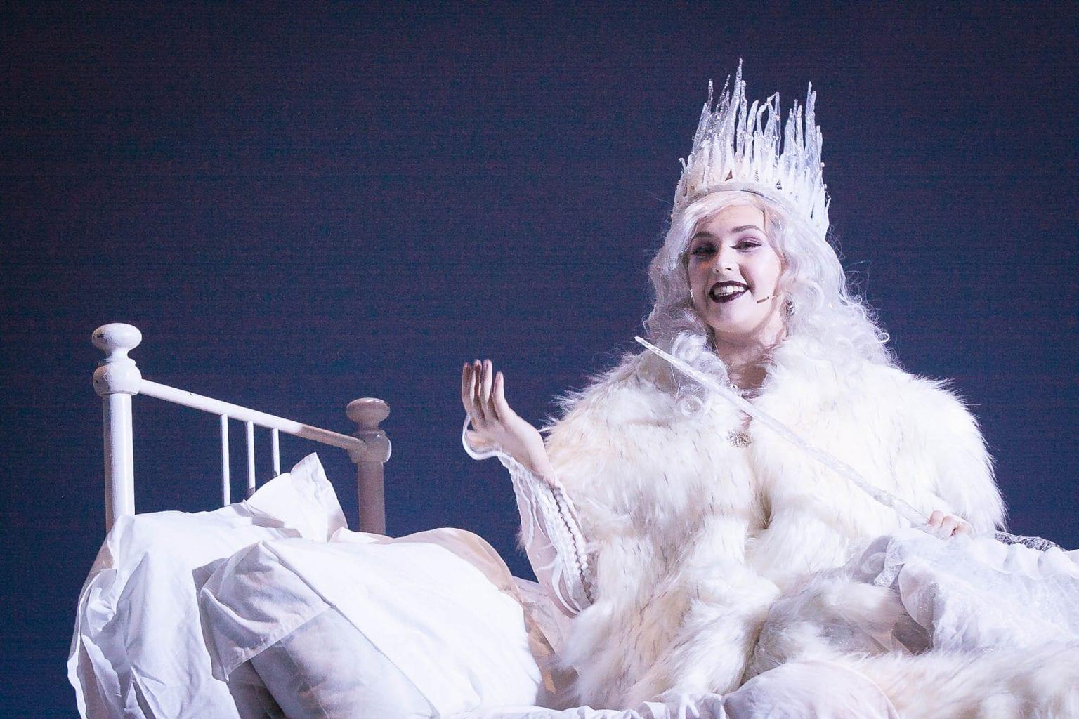 A person in costume as the White Witch from the Chronicles of Narnia sits on a bed.