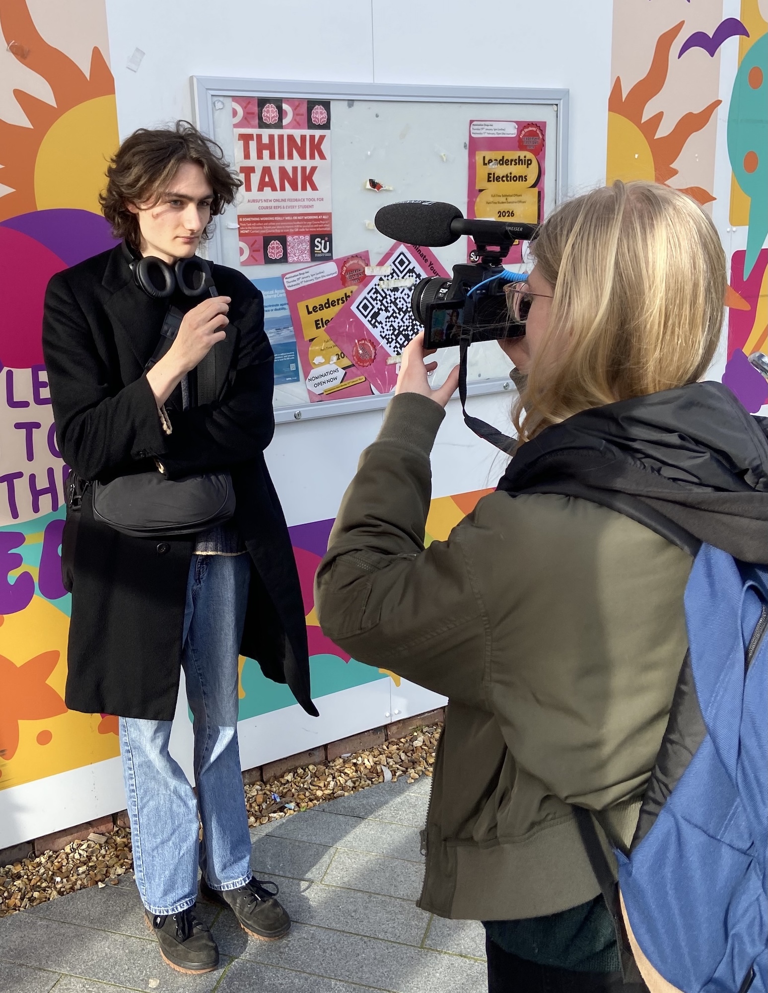 Young person being interviewed by BA (Hons) Film Production student against colourful wall mural with "THINK TANK" poster and geometric patterns.
