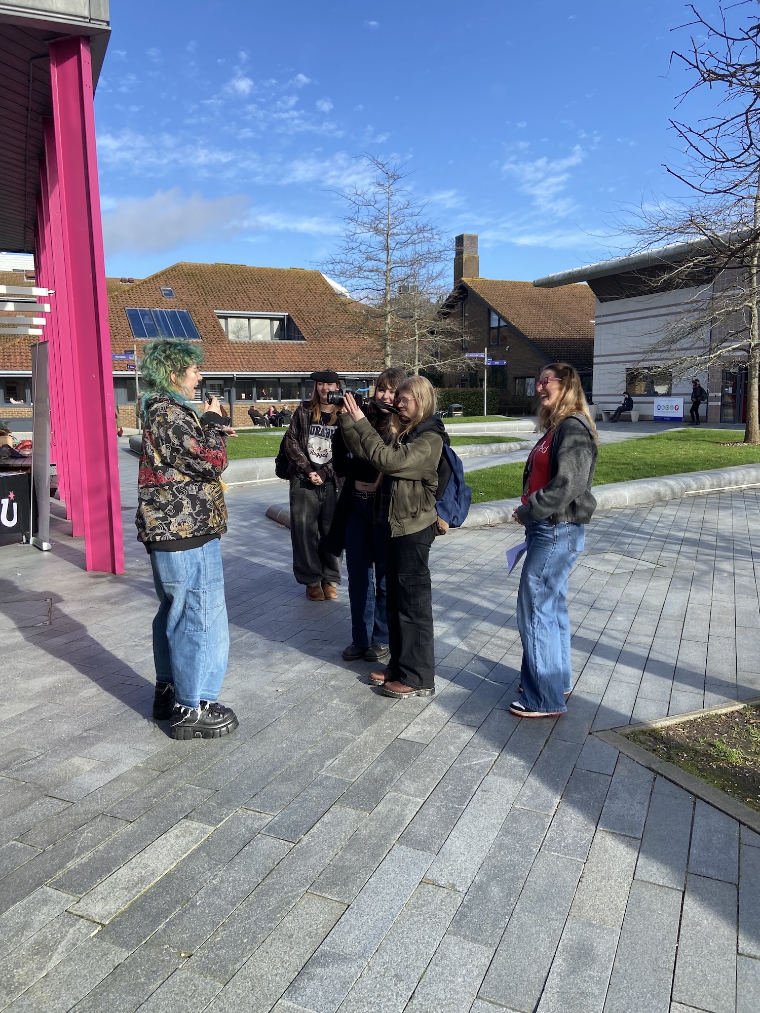 Five AUB students standing on grey paved area outside modern buildings with brown roofs. Pink structure visible on left, blue sky above.