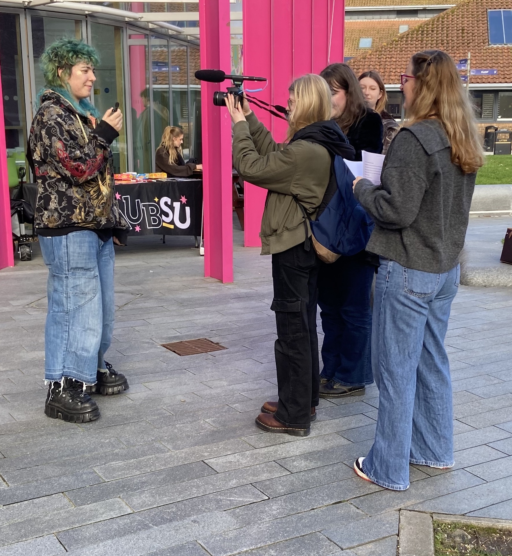 Person with green hair in floral jacket stands near bright pink building entrance whilst group films with camera on courtyard paving.