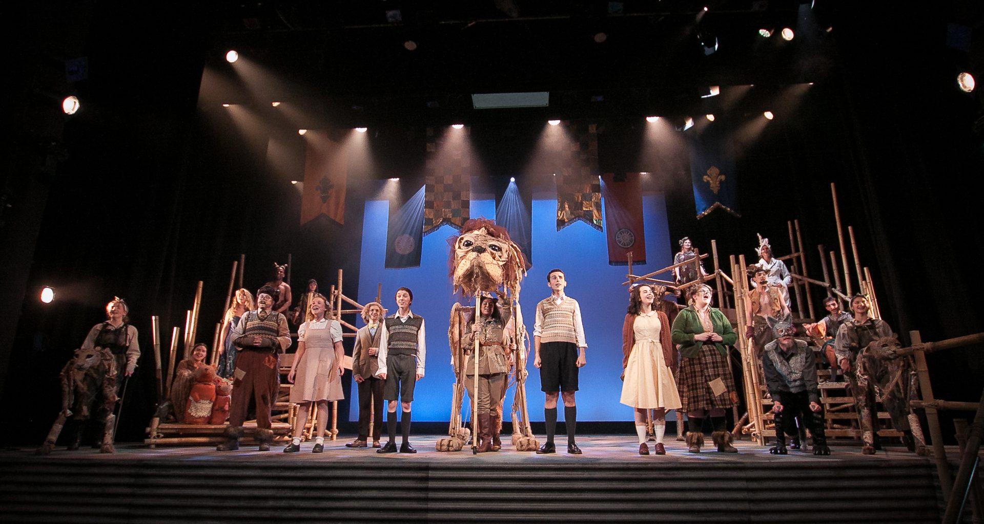 A group of actors in costume stand mid-song on a stage. Several tapestries hang around the spotlights. A large lion puppet is in the middle of the stage.