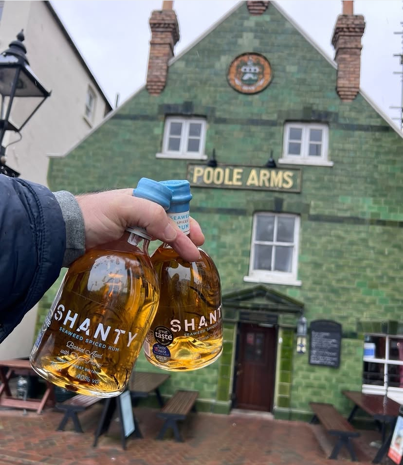Hand holding two Shanty bottles of brown rum outside the green-tiled Poole Arms pub.