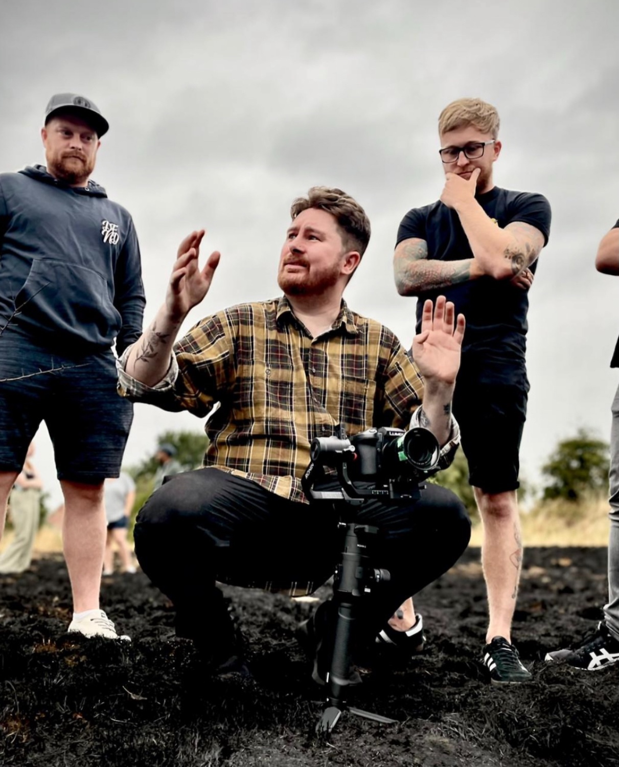 Three men, including filmmaker J White, outdoors with camera equipment. Centre man in checked shirt gesturing with hands raised, flanked by two others standing.