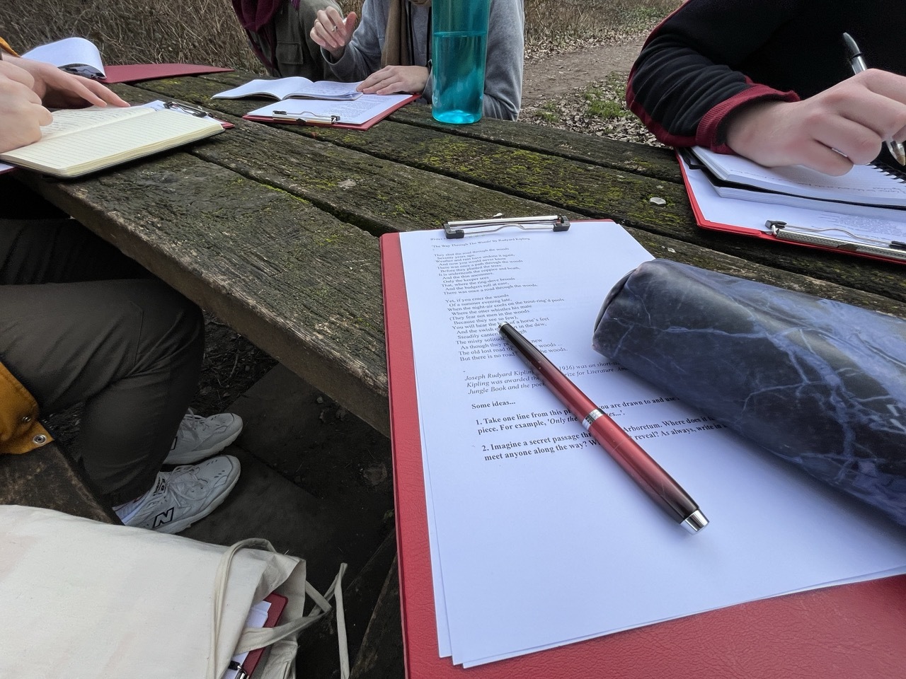 Red clipboards with text on a mossy wooden table outdoors, people writing.
