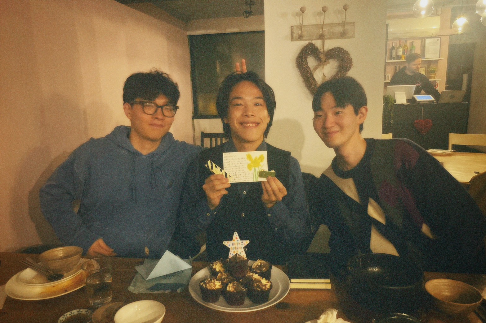 Three Asian students sat on a table indoors with happy birthday cupcakes, Seiji in the middle smiling with a flower card.