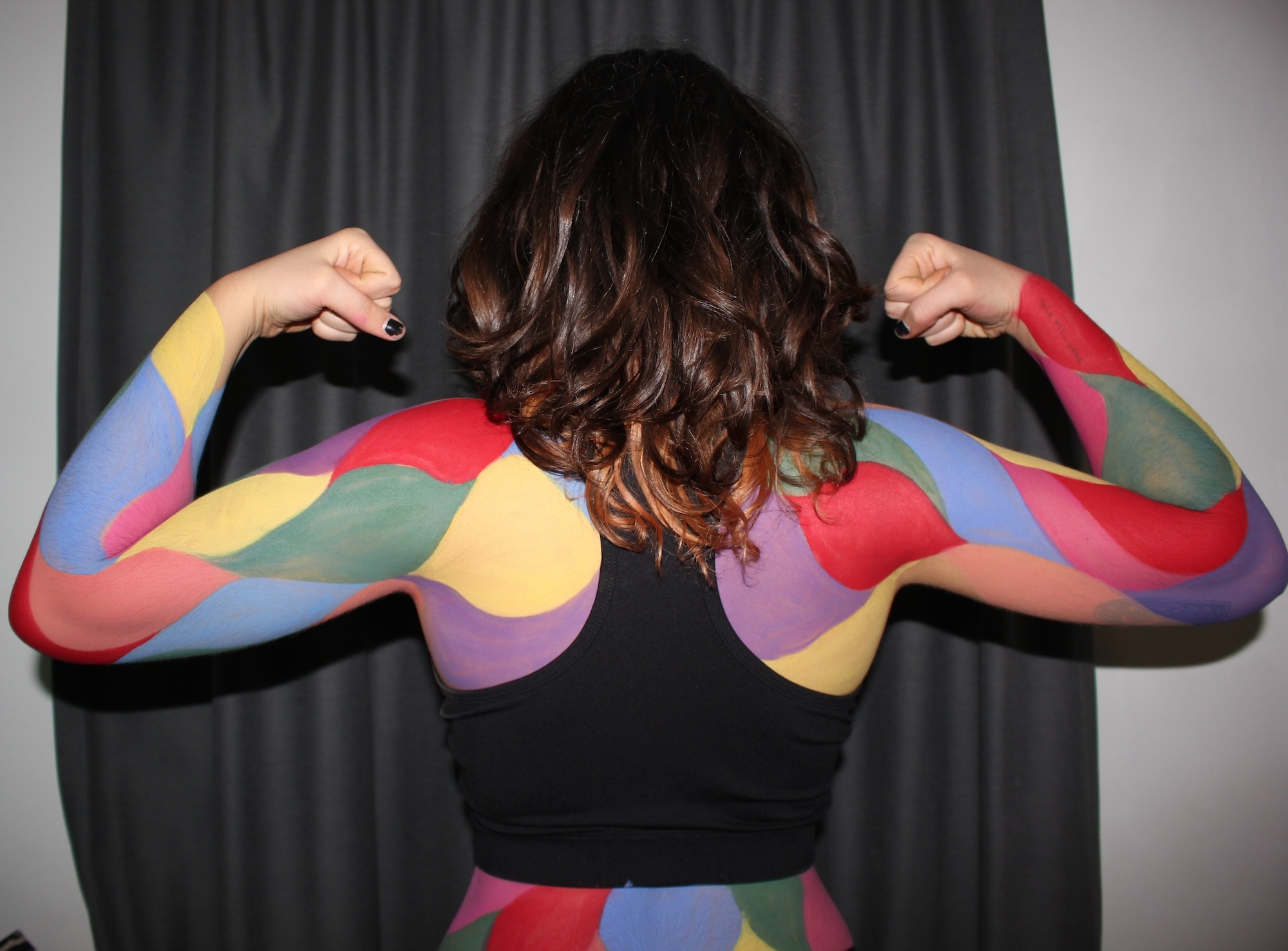 A young person viewed from the back, showing their arms, shoulders and waist painted with a multi-coloured harlequin pattern.