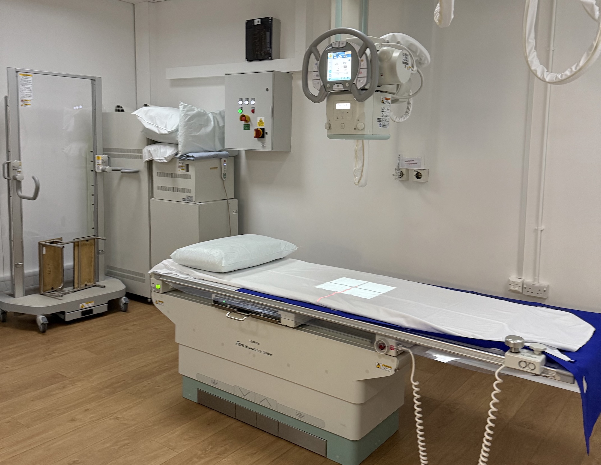 A medical examination room featuring an examination bed with a white sheet and pillow. To the left, there is a cabinet with medical equipment, and a machine on the ceiling with a control panel is visible above the bed.