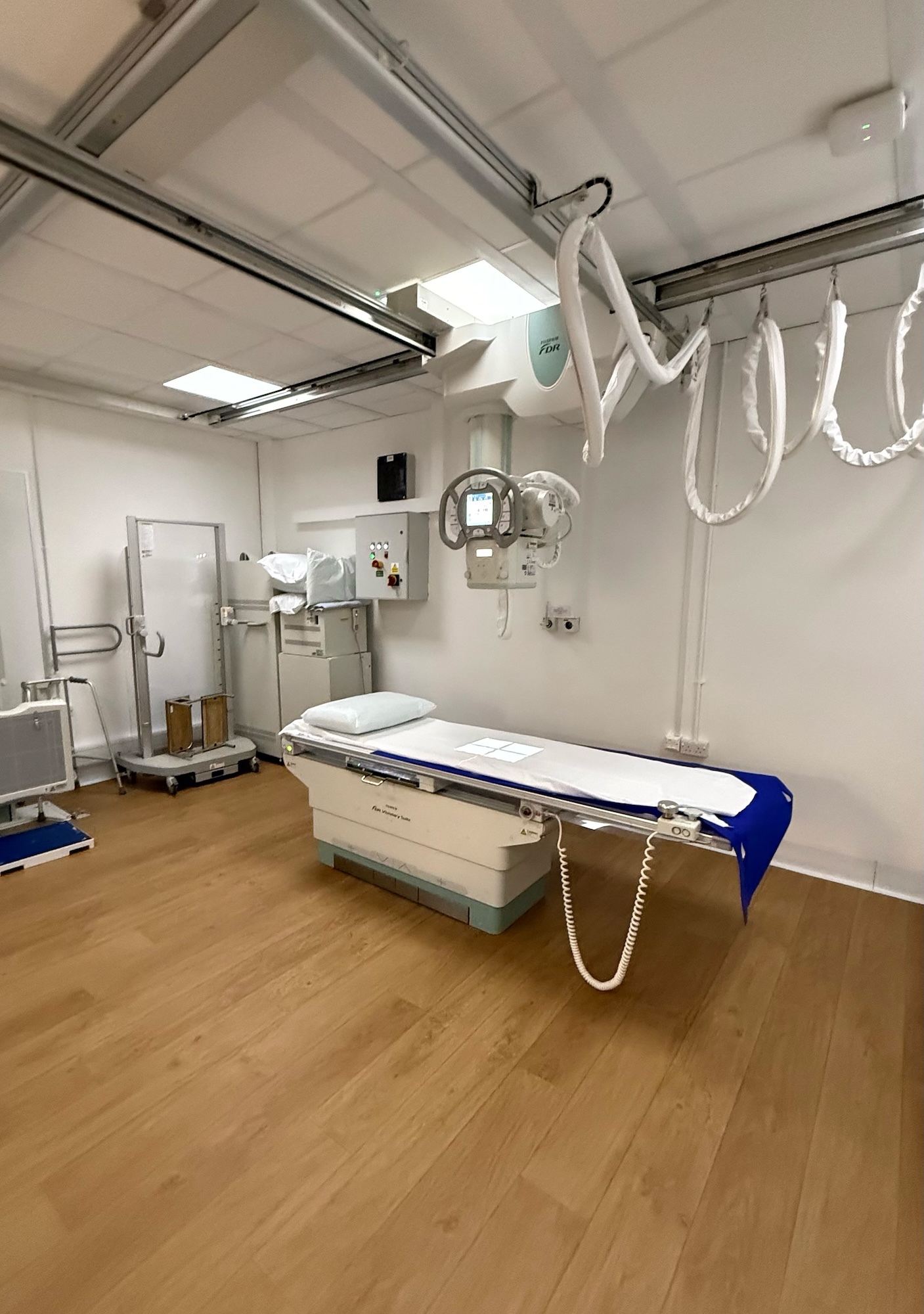 Medical examination room with white walls, ceiling-mounted X-ray equipment, examination table with blue fabric, wooden flooring, and door.