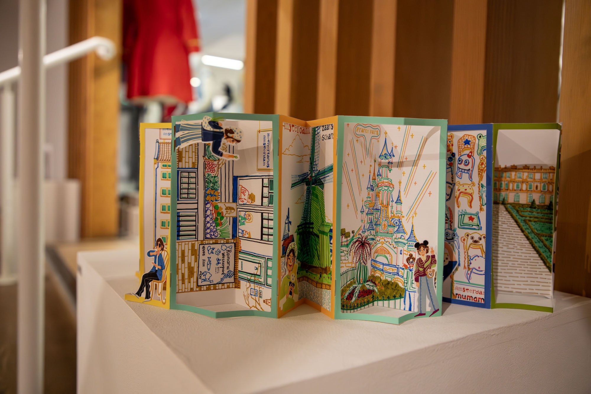 A leaflet partly unfolded on display to show several colourful illustrations, scenes including characters in front of a windmill and the castle at Disneyland.