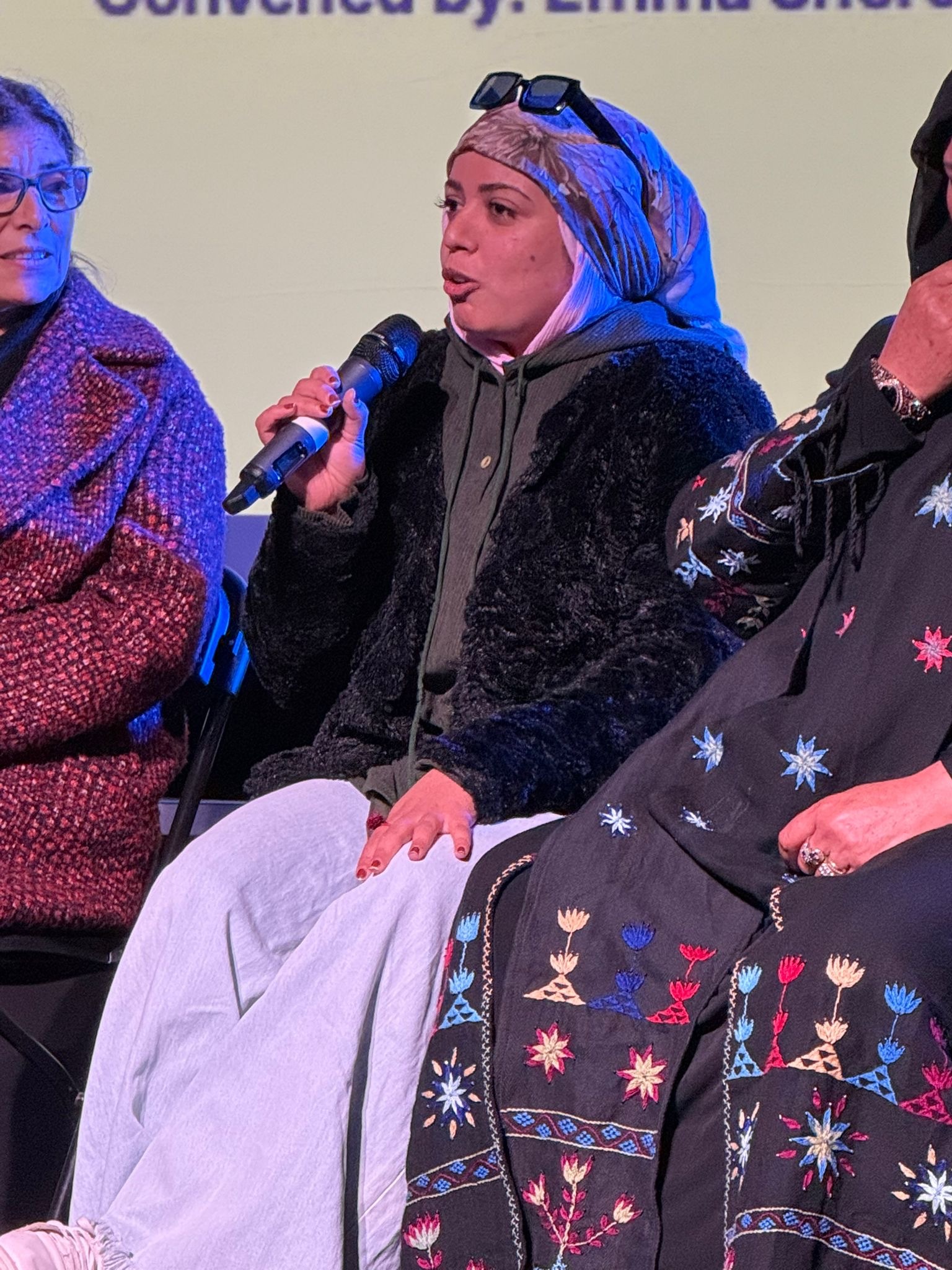 Woman in blue headscarf speaks into microphone at panel discussion, flanked by two other seated participants under stage lighting.