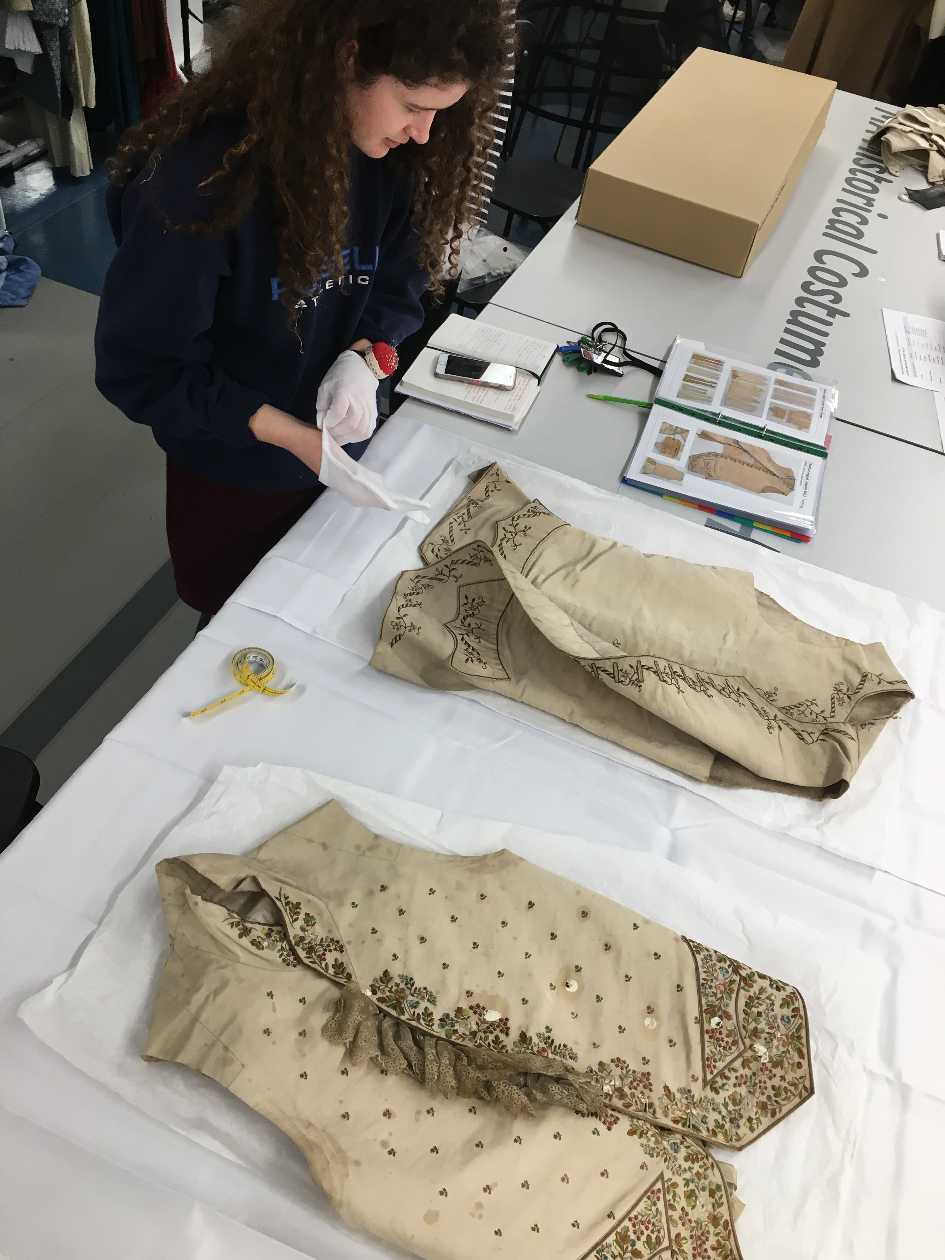 Student working on a pair of historical-style waistcoats lying on a long table.