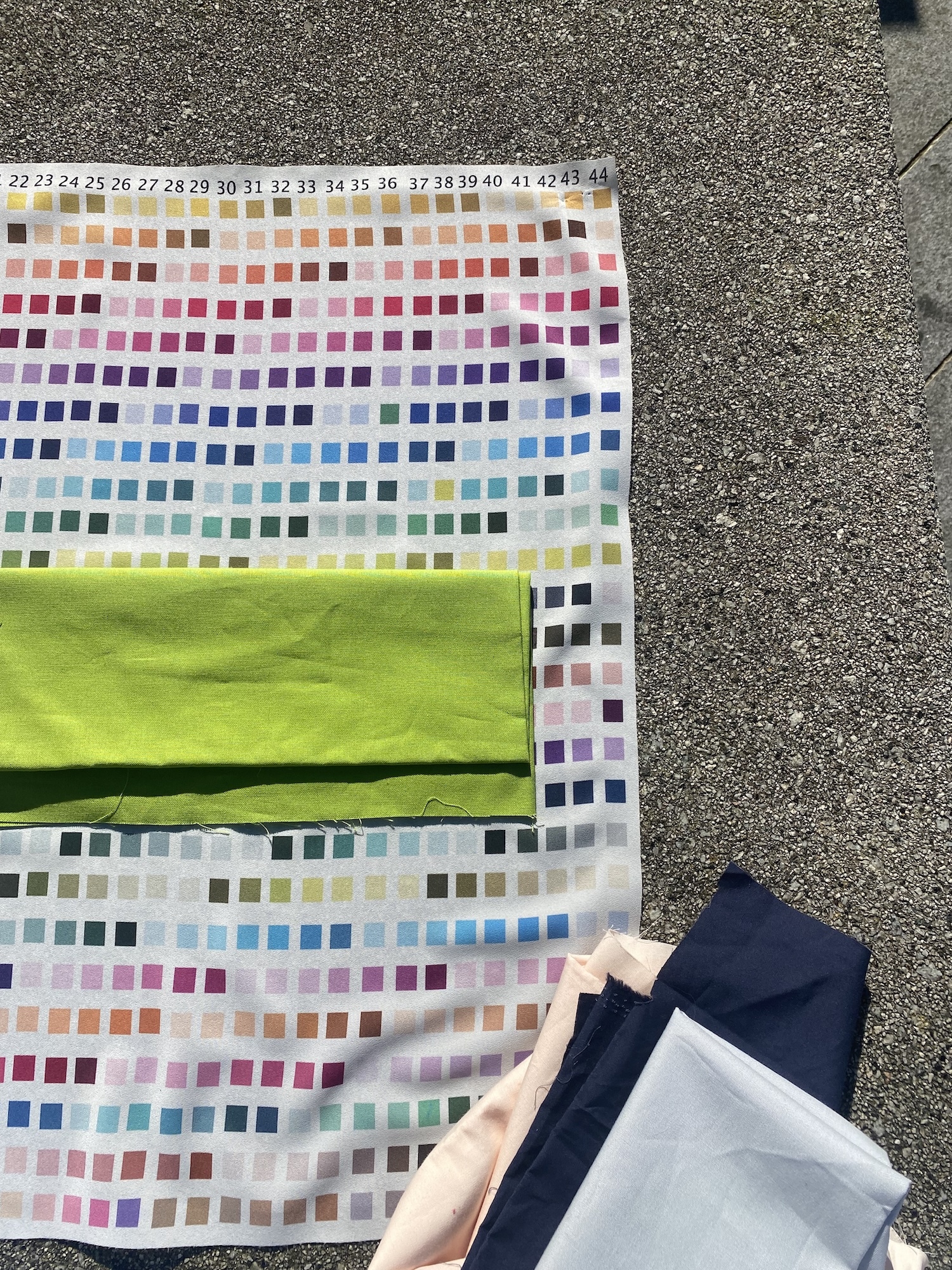 Colour chart with numbered squares in rows from orange to purple hues, bright green fabric sample across middle, on tarmac surface.