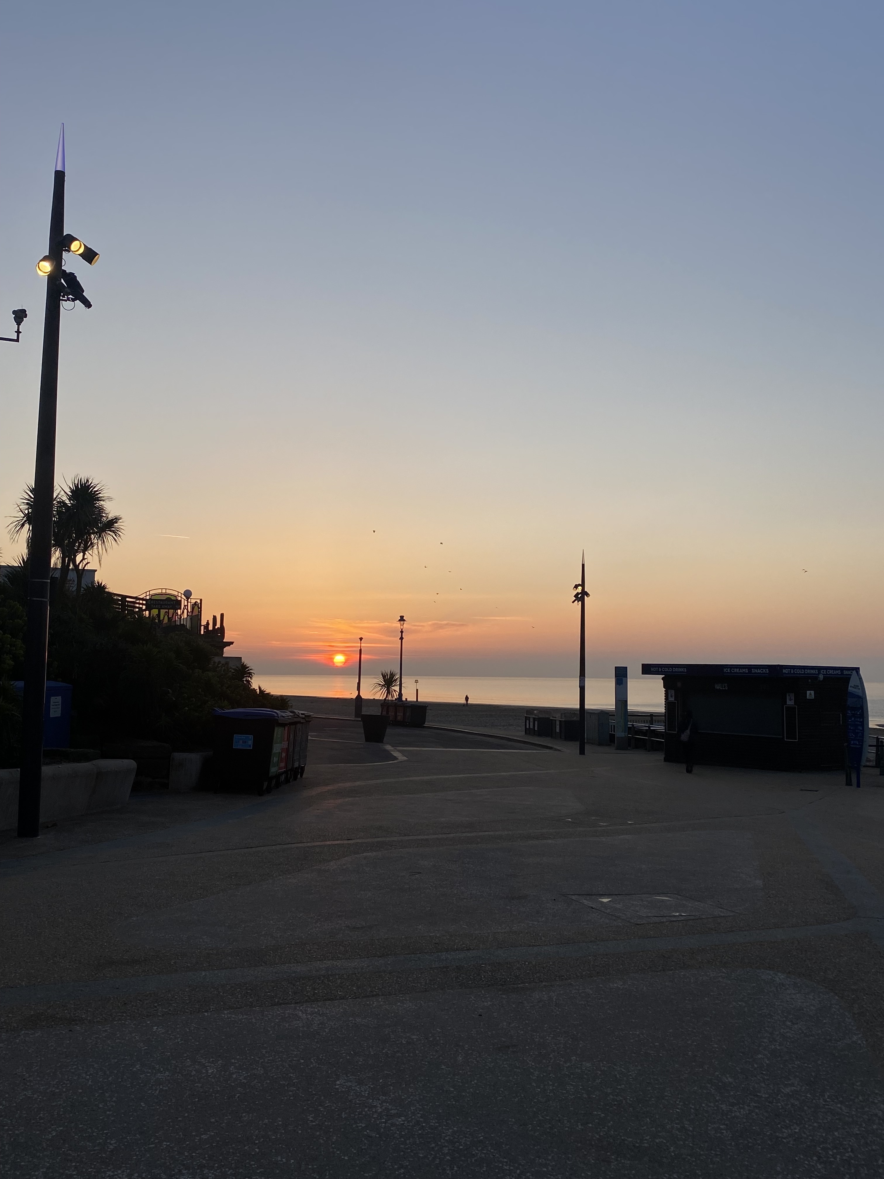 Sunset over seaside promenade with lamp posts, palm trees, and buildings silhouetted against orange and blue gradient sky.