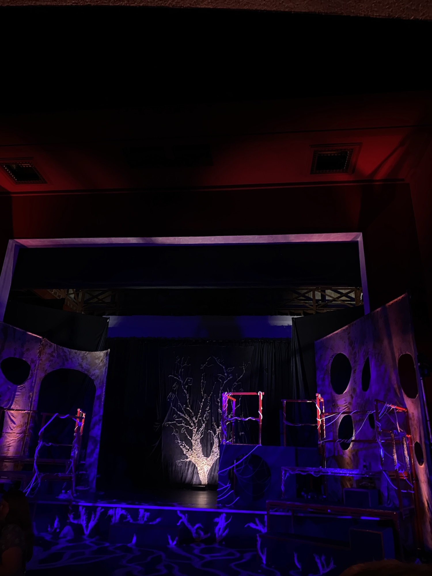 A stage setting featuring an abstract backdrop with a white tree design, illuminated by purple and red lighting. The stage has structures that appear like scaffolding and circular cut-outs, with a dark curtain in the centre.