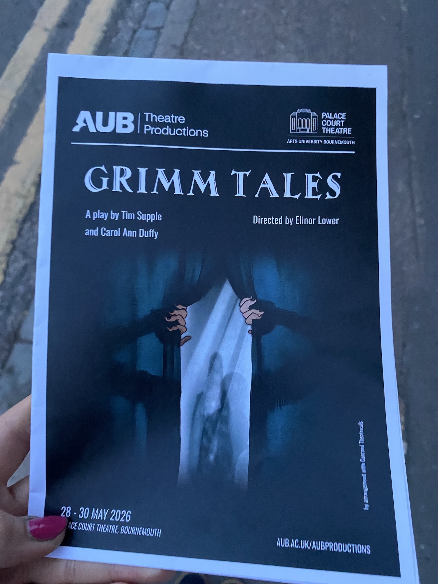 A hand holding a promotional flyer for a theatre production titled 'Grimm Tales'. The flyer features dark imagery, a pair of hands pulling back a curtain, and details about the production, including the play's authors, director, and performance dates.