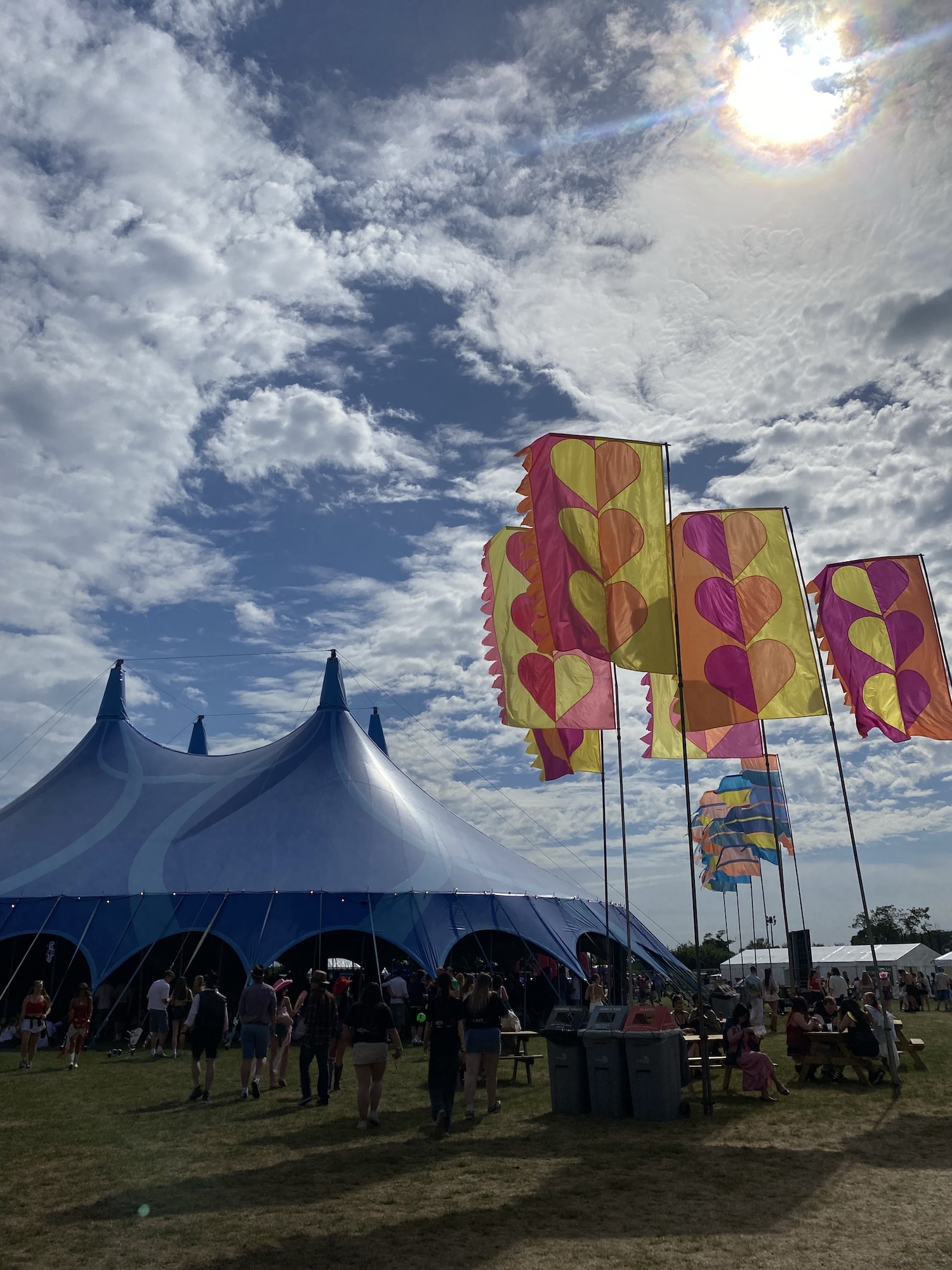 A large blue event tent stands on a grassy field beside tall colourful flags, with people gathered underneath a cloudy sky and bright sun.
