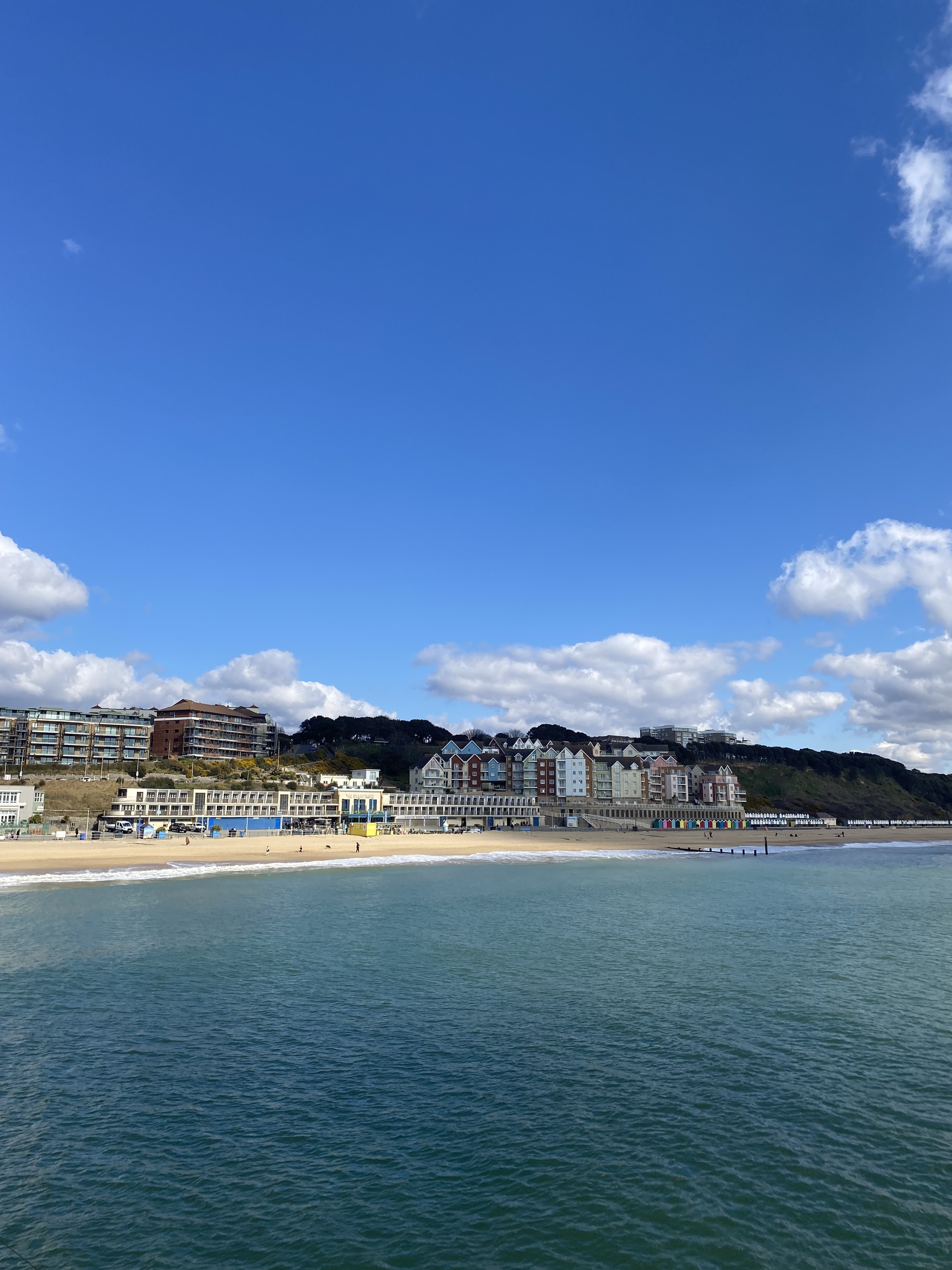 Coastal town with multi-storey buildings along seafront, sandy beach, calm turquoise water, white clouds against bright blue sky.