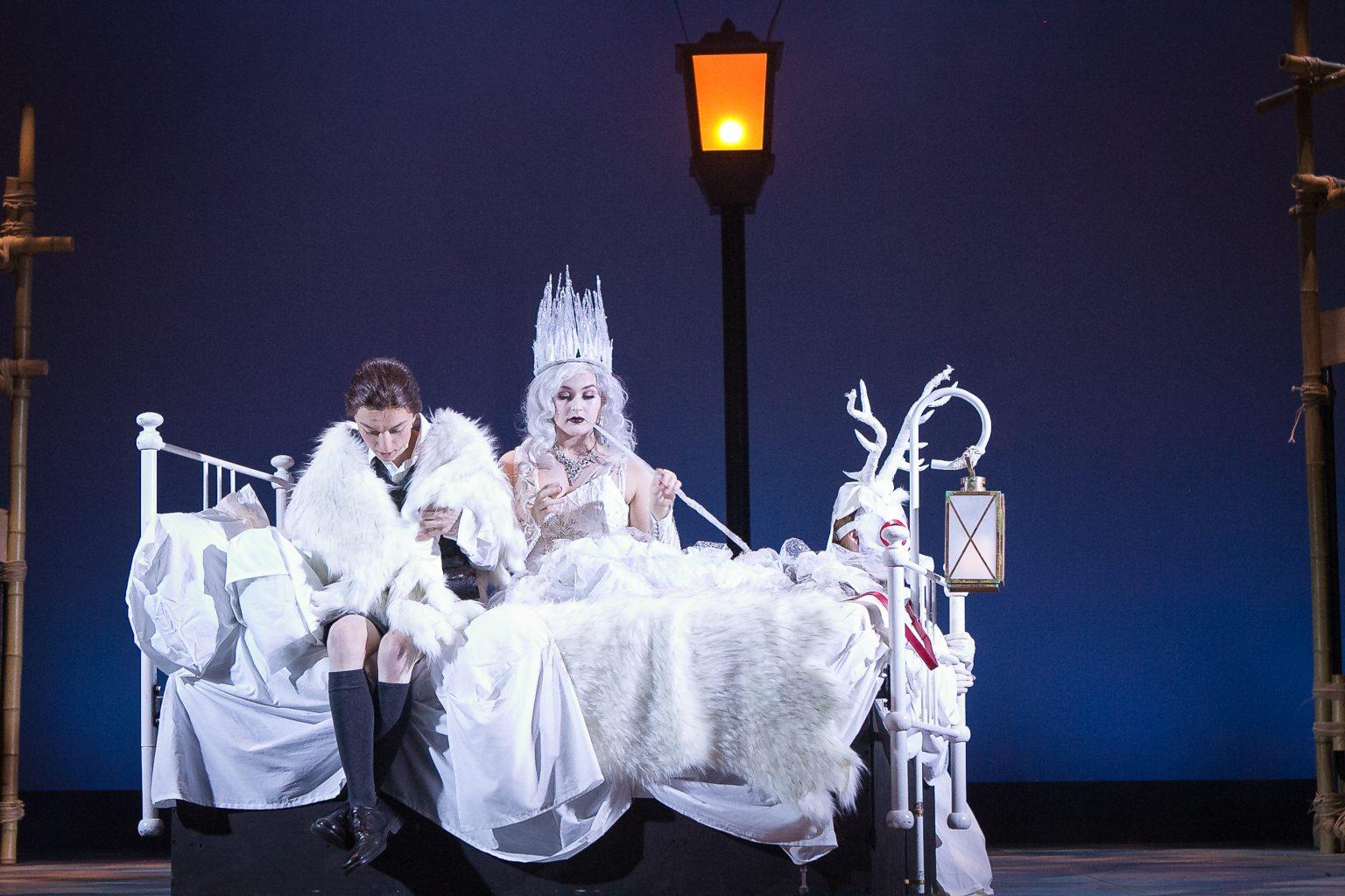 Two actors in costume - one as a white witch with an ice crown, the other as a school child with a fur shawl wrapped around them - sitting on a bed.