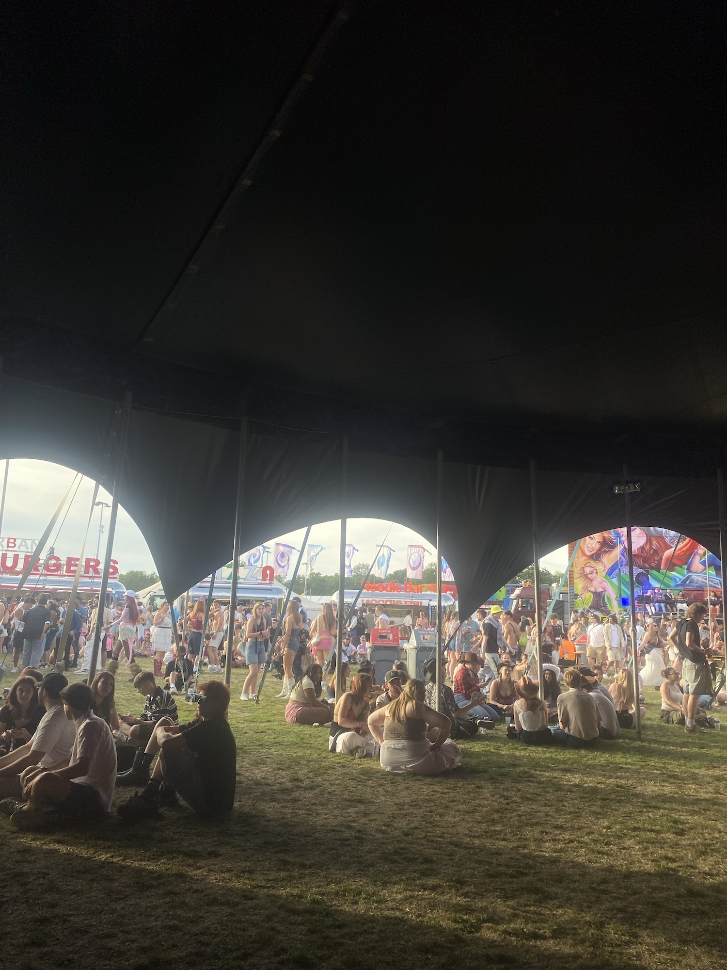People sitting on grass under a large dark canopy at an outdoor festival, with fairground rides, stalls and crowds visible beyond the open side.