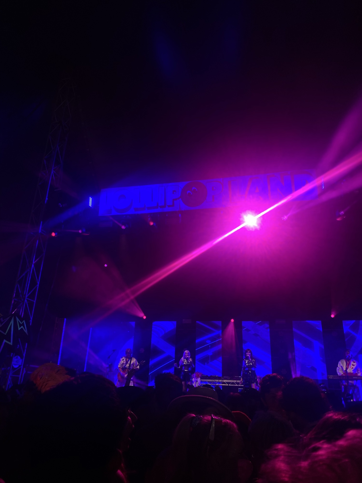 A live band performs on a stage lit with purple and blue lights, with a crowd in silhouette in the foreground.