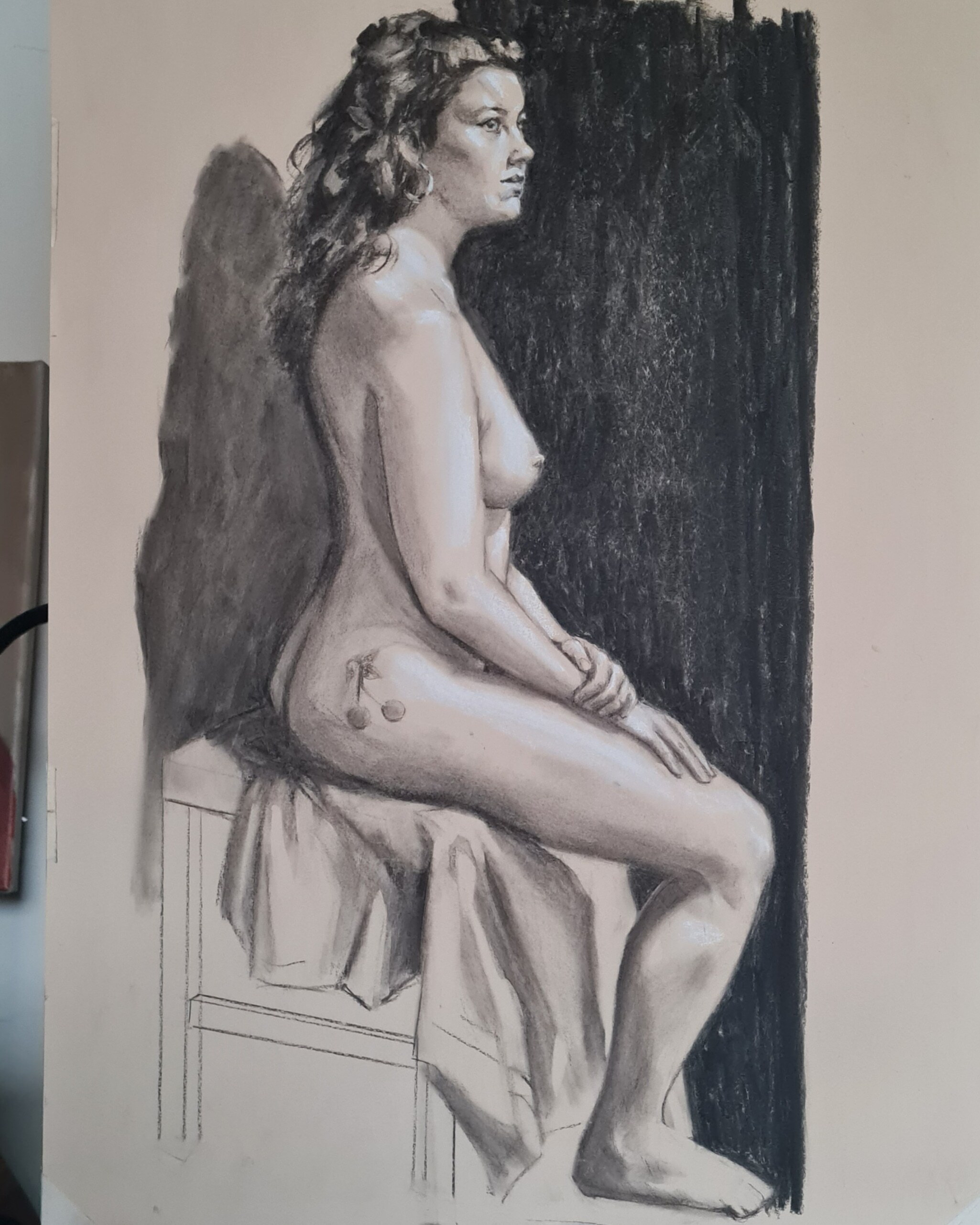 Nude figure drawing in charcoal and pastels showing seated woman with curly hair against dark background, pale flesh tones on cream paper.