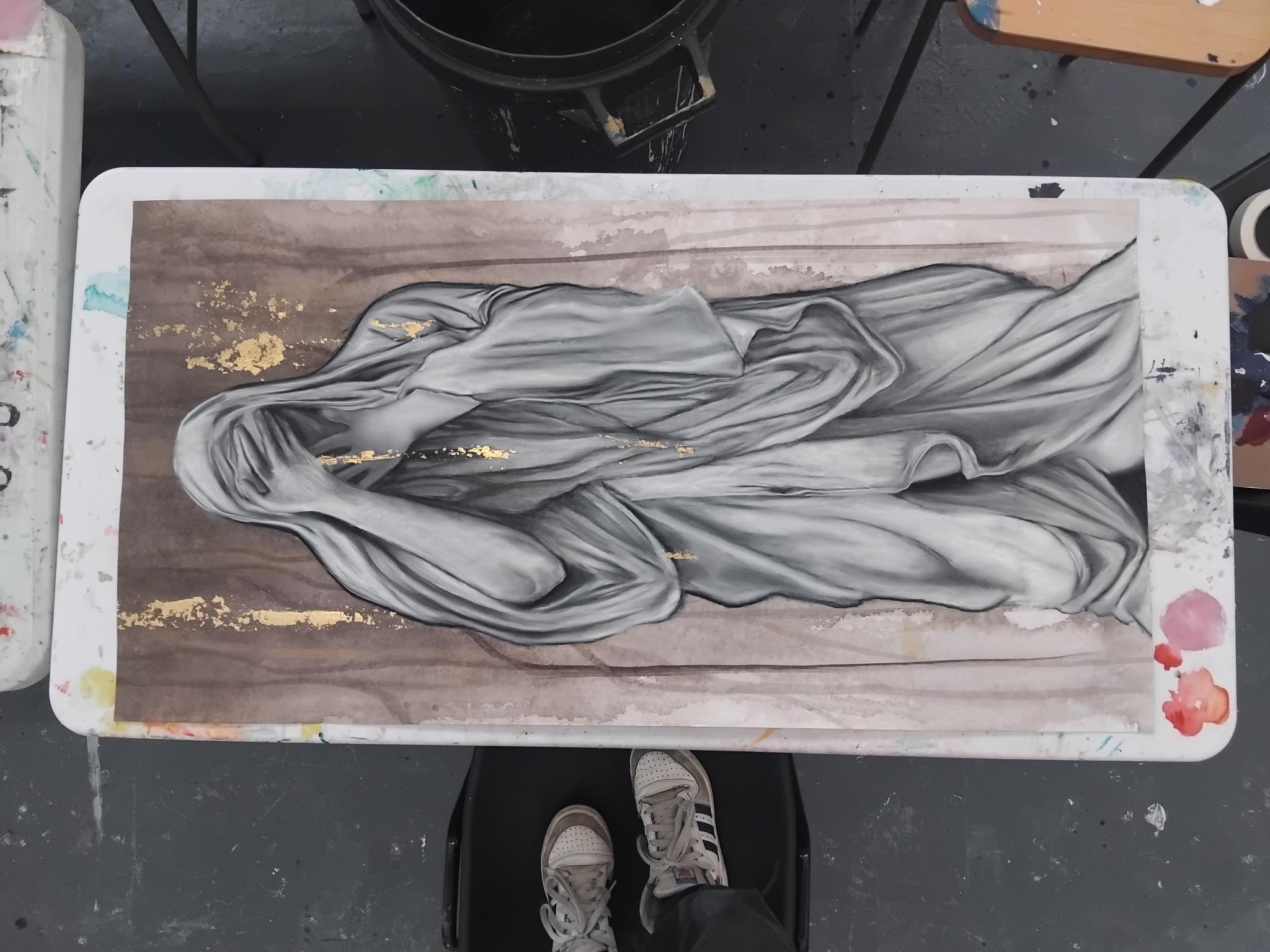 A large artwork featuring a draped figure in grey tones set against a wooden background, with subtle gold accents. The painting is displayed on a table, with feet visible at the bottom. The surrounding area includes furniture items and a dark bucket.