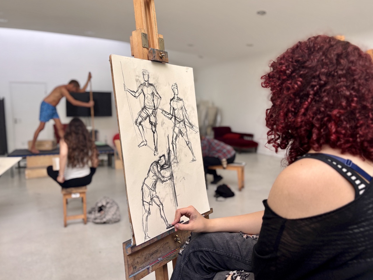 A woman with curly red hair sketches a figure on an easel in a bright studio, with an artist posing in the background and sketch studies on the canvas.