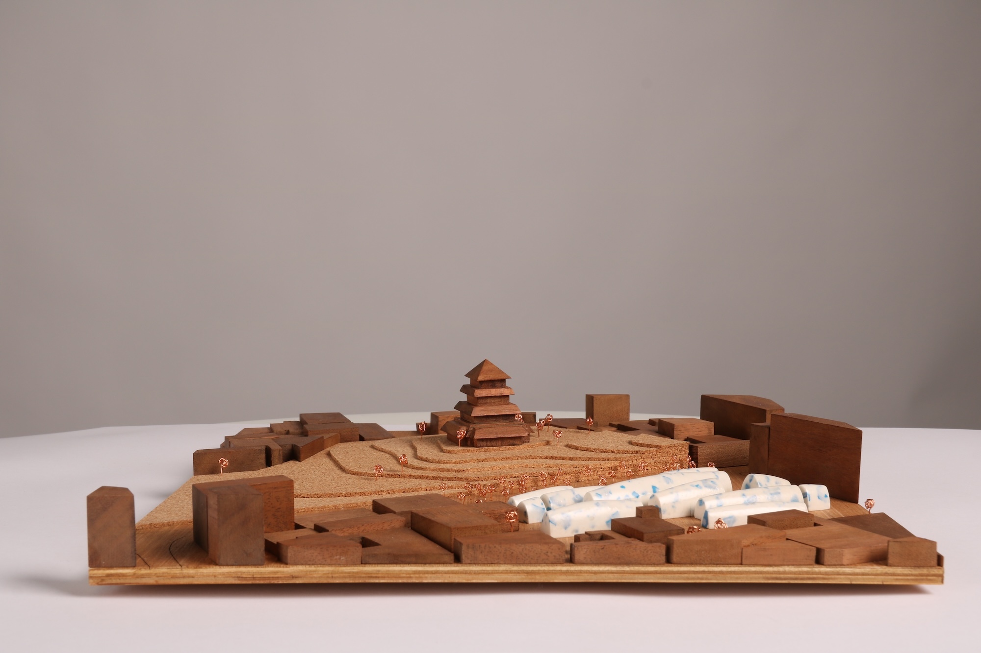 Wooden architectural model with tiered central structure and surrounding buildings on white surface against grey background.