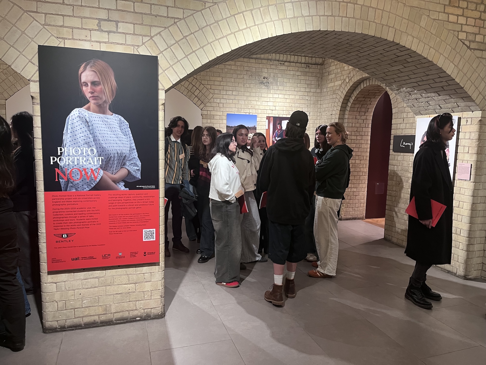Visitors stand in a tiled gallery corridor beside a large poster for Photo Portrait Now, with arched brick ceilings and framed artworks in the background.